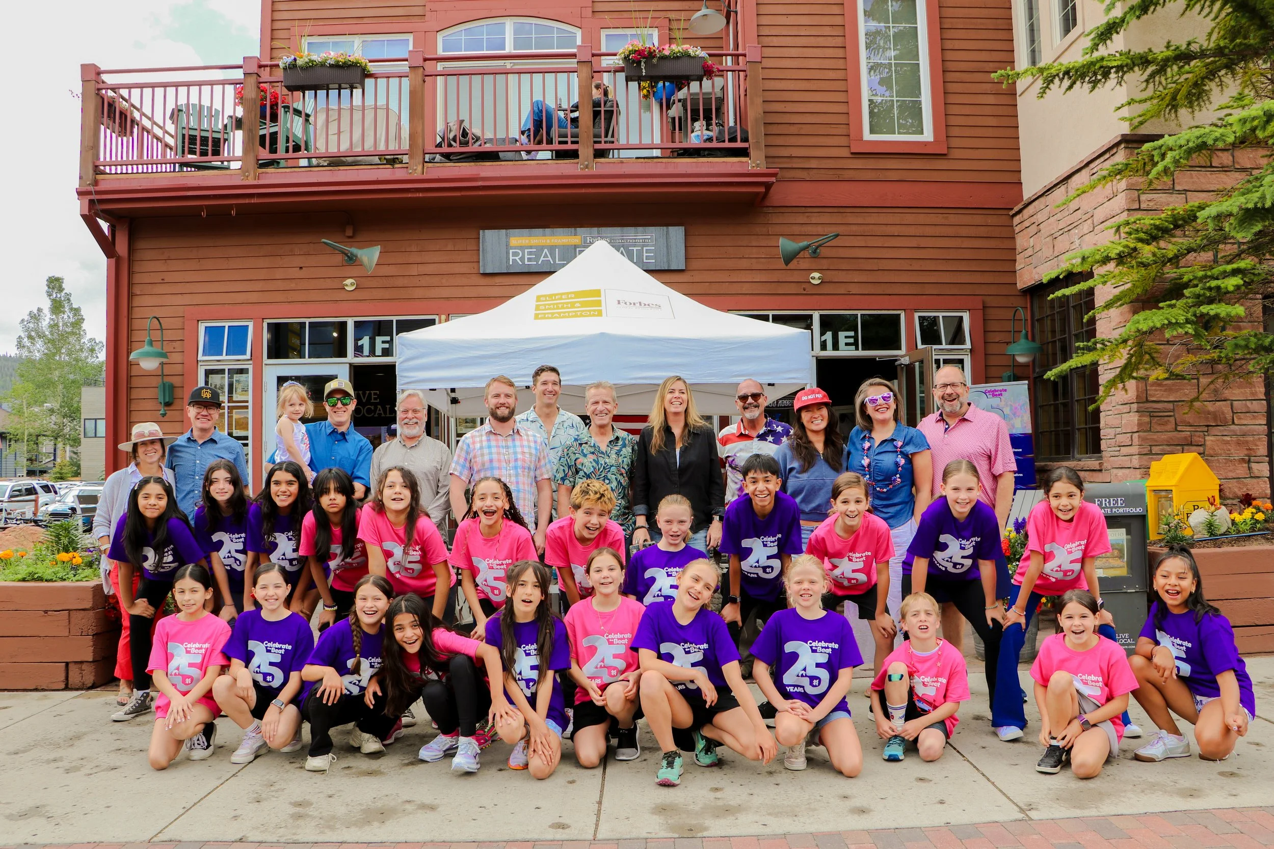 A large group of children and adults posing for a photo outside of a red building with a balcony. The children are wearing colorful shirts, including purple and pink, with some shirts celebrating 25 years. The adults stand behind the children, smilin