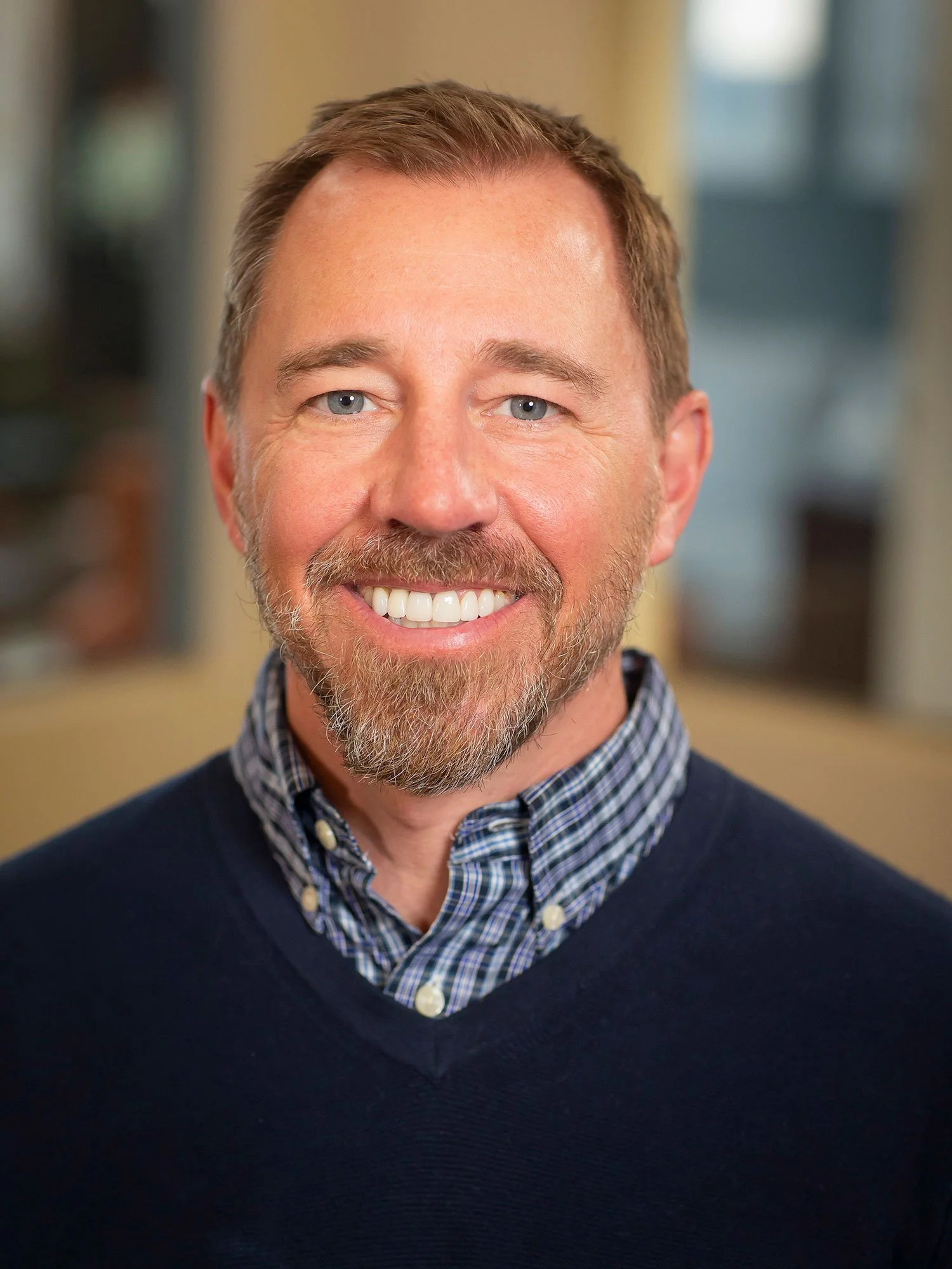 A smiling man with blue eyes, light brown hair, and a beard, wearing a dark blue sweater over a collared shirt with a checkered pattern, standing indoors with a blurred background.