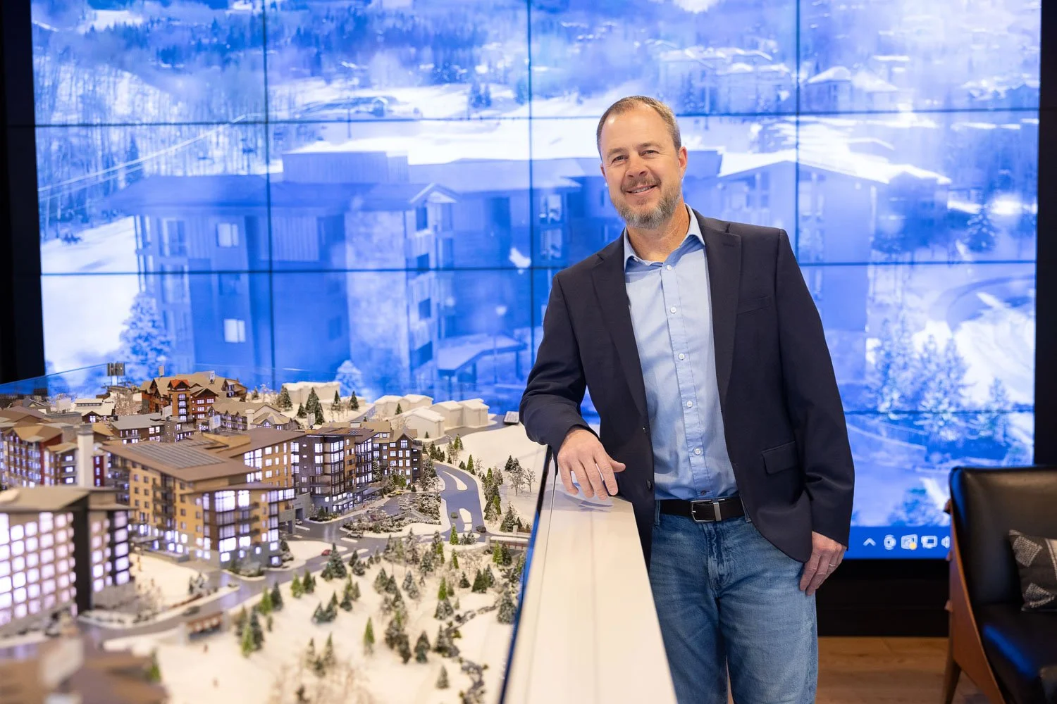 A man in a suit jacket and jeans standing beside a detailed architectural model of a residential complex, with a large digital screen showing a winter scene in the background.