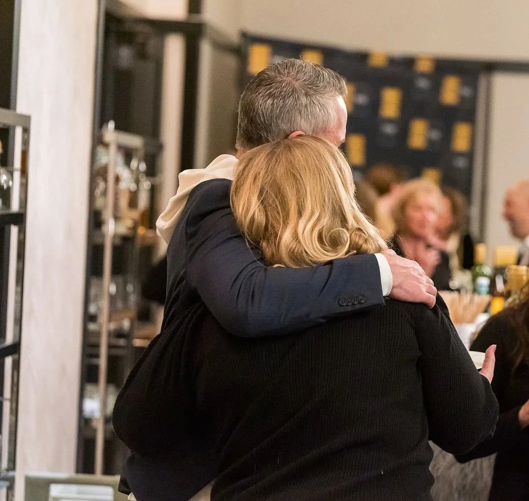 Two people sharing an embrace at a social gathering or event, with other people in the background.