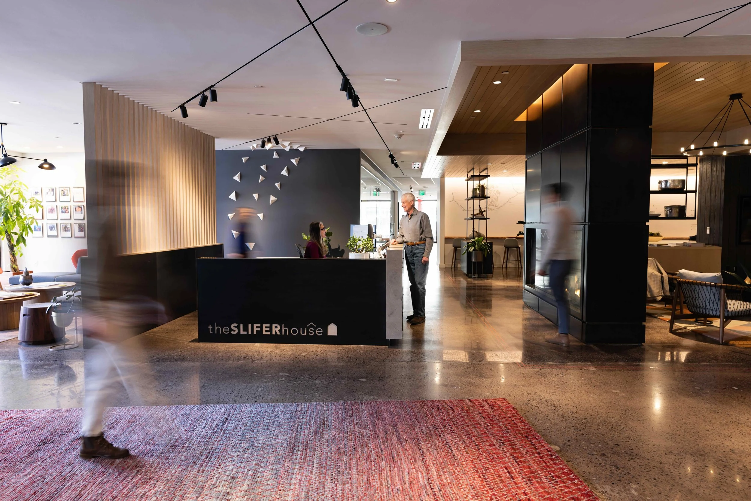 Modern hotel lobby reception area with a black counter displaying 'theSLIFERhouse' logo, a man and a woman interacting, contemporary lighting, and stylish decor including a wall with floating geometric shapes and a door in the background.