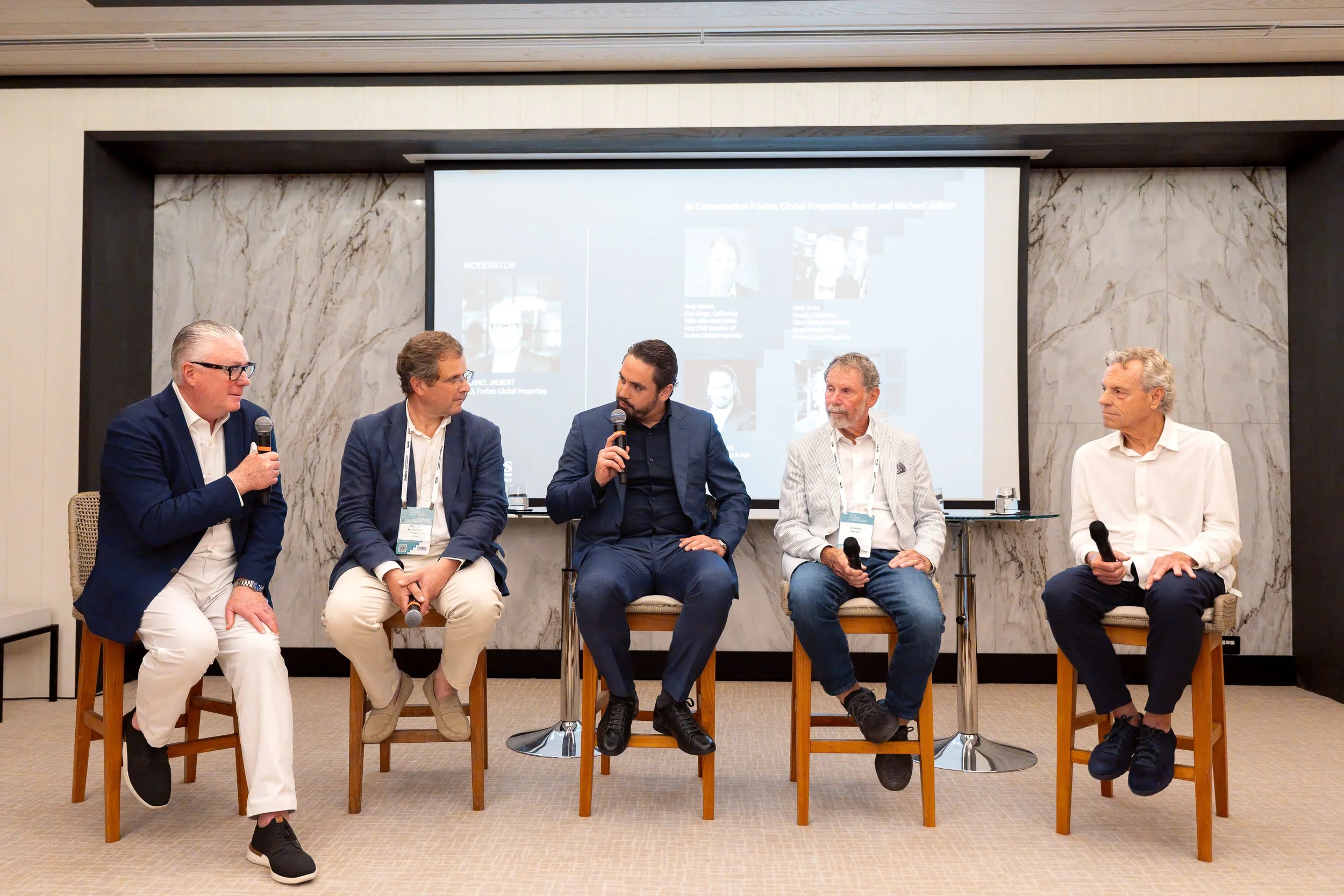 Five men sitting on chairs with one holding a microphone, participating in a panel discussion in front of a large screen displaying a presentation. The background features a marble wall.