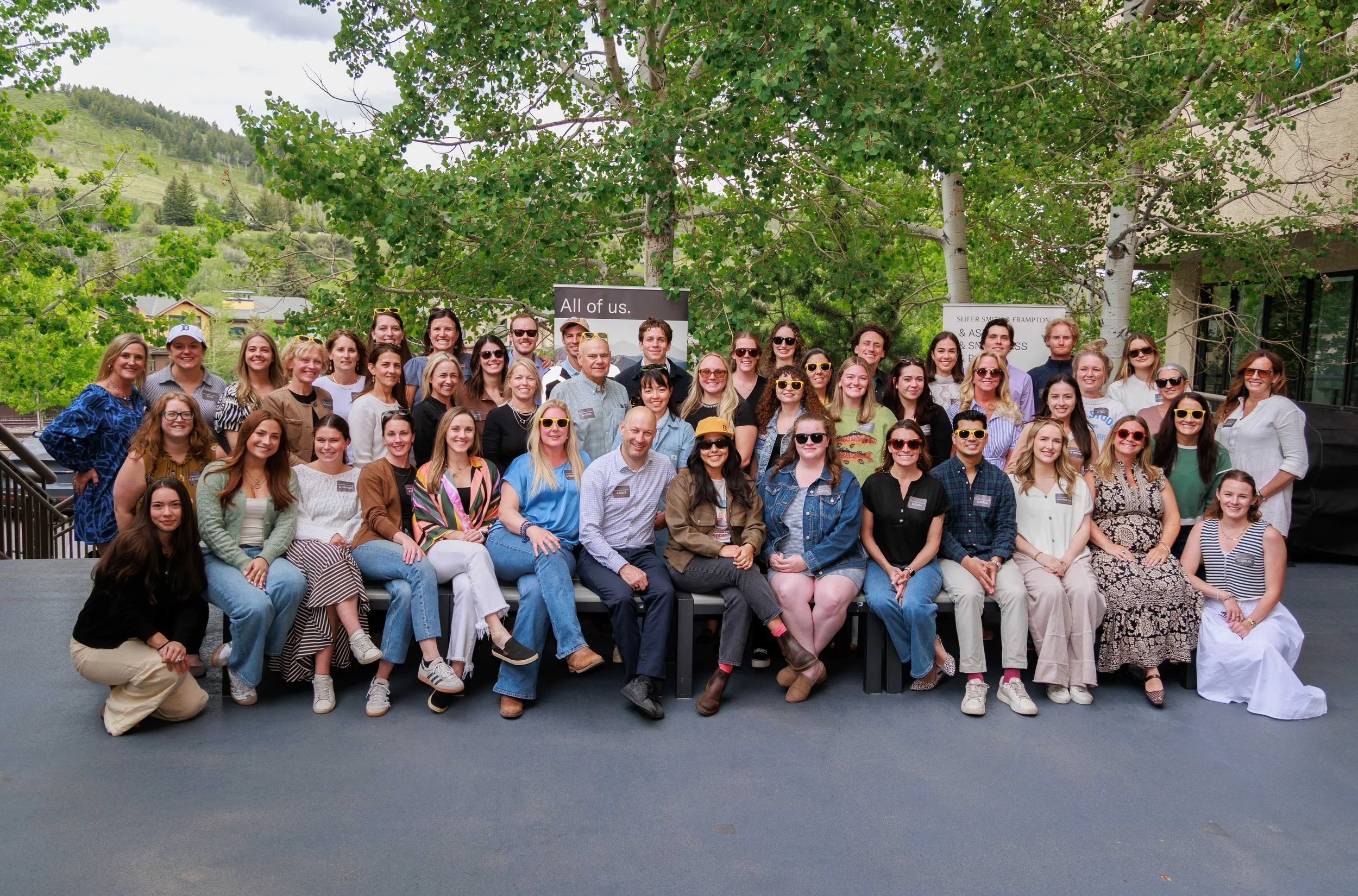 Large group of people posing outdoors under trees with a hillside in the background, some individuals wearing sunglasses and smiling.
