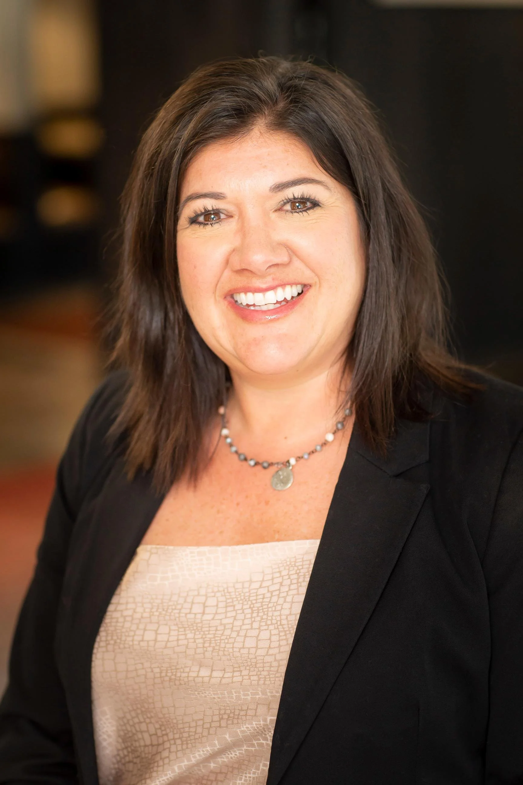 Close-up of a smiling woman with dark brown hair in a professional setting, wearing a black blazer, a beige textured top, a string of beads, and a pendant necklace.