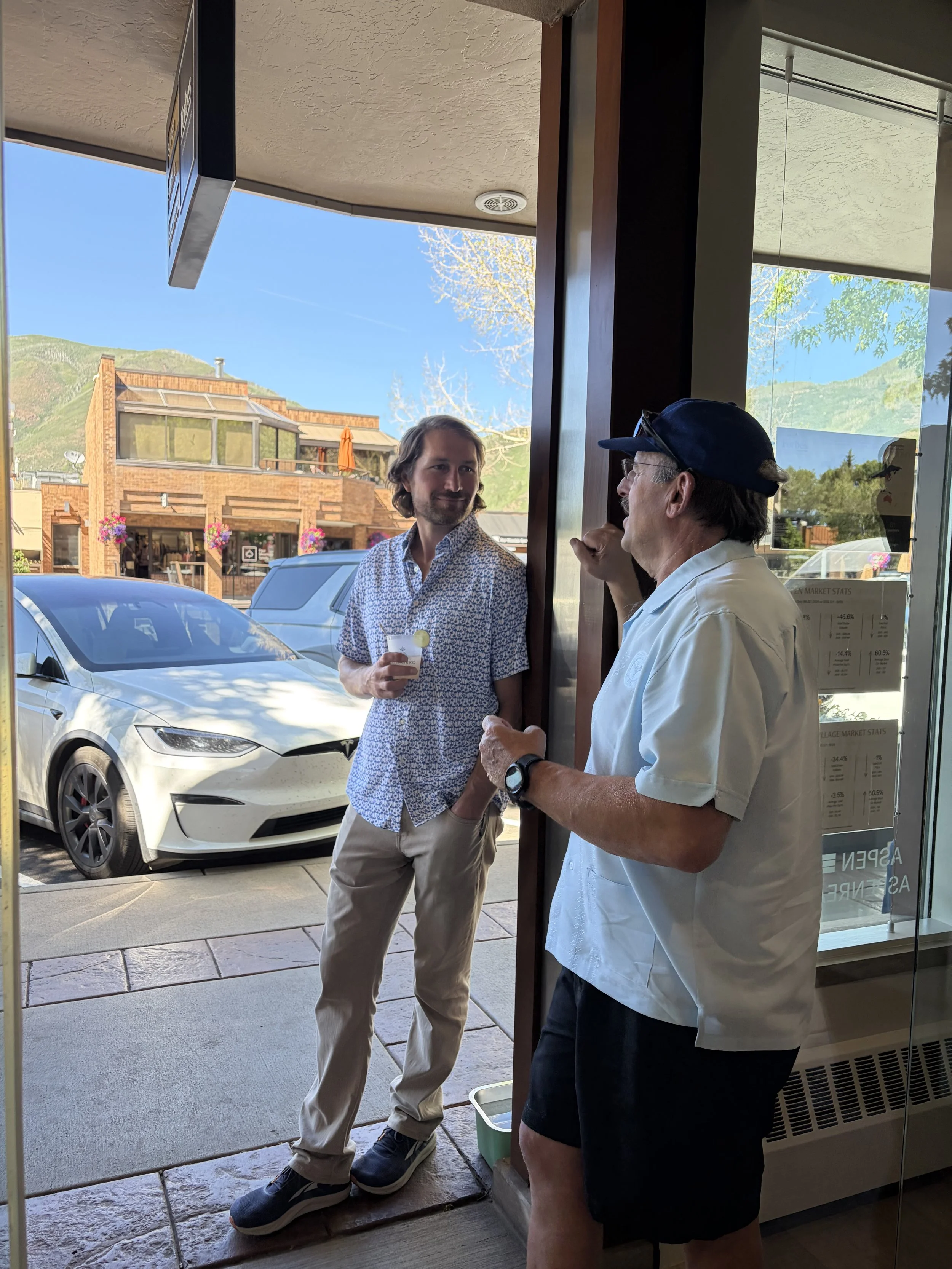 A man with a beard and a patterned shirt holding a cup, talking to another man with glasses, a cap, white shirt, and black shorts, inside a cafe or restaurant near the entrance, with cars outside and a mountainous landscape in the background.
