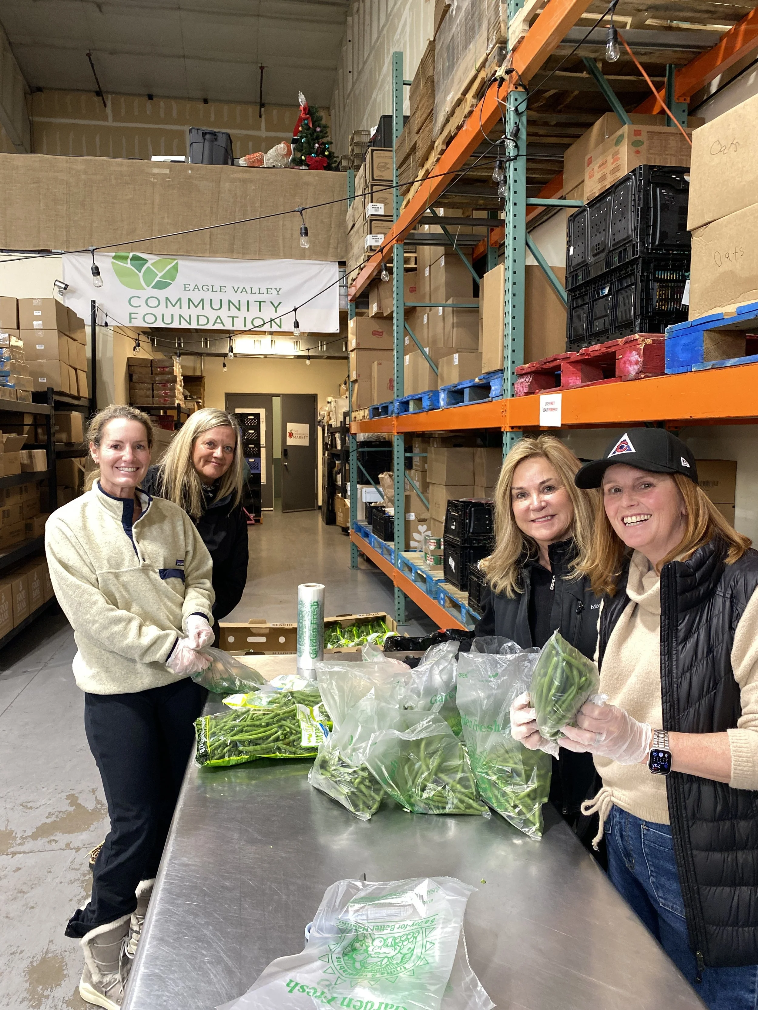 Four women standing in a warehouse, packing fresh vegetables into plastic bags, with shelves of boxes and a small Christmas tree in the background, and a banner that says 'Eagle Valley Community Foundation'.