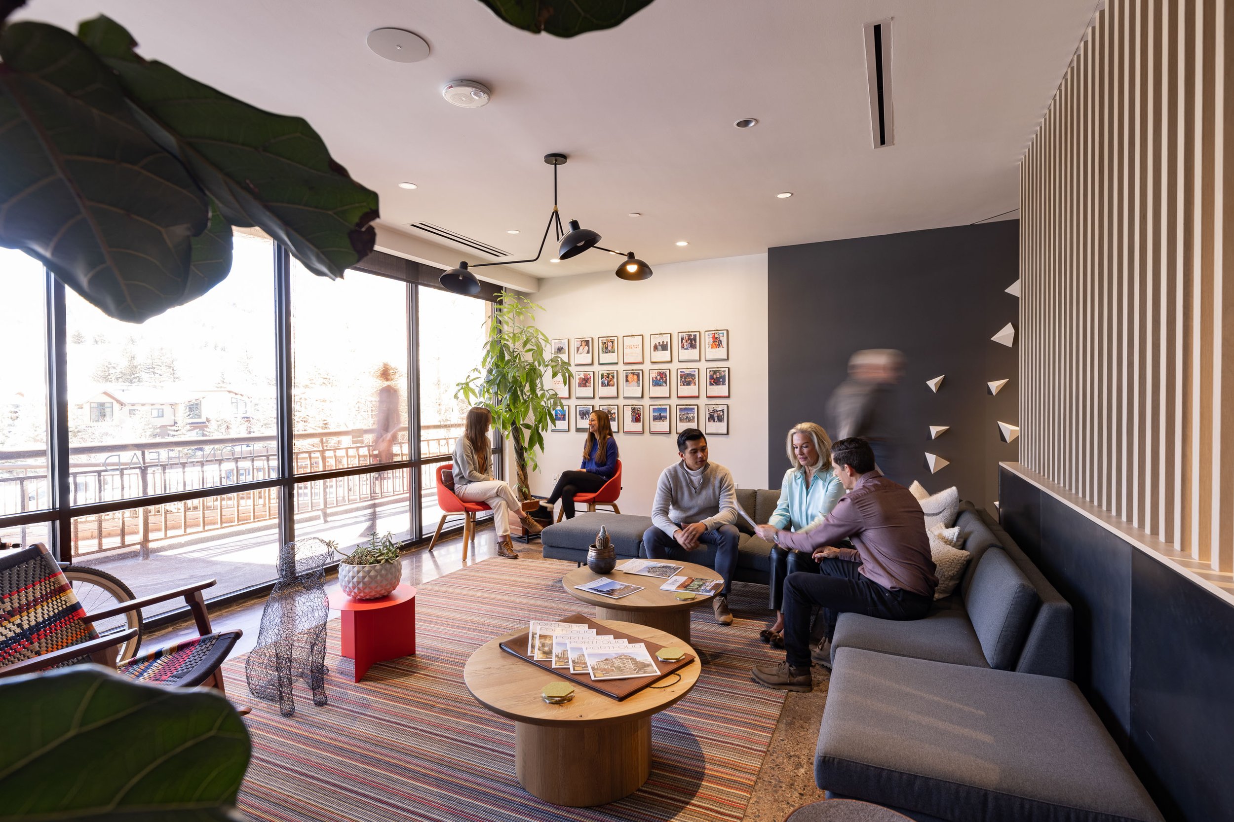 People sitting and chatting in a modern lounge area with large windows, framed photos on the wall, and various plants.