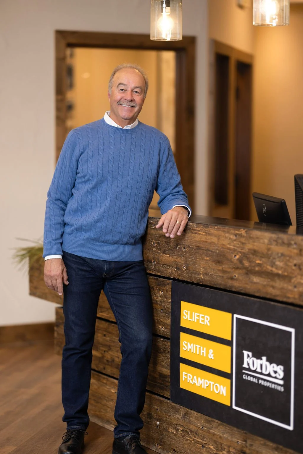 A smiling man in a blue sweater and jeans standing at a reception desk made of wood. Behind him is a wall with warm lighting, and the desk displays a sign for Forbes listing SLIFER, SMITH & FRAMPTON.