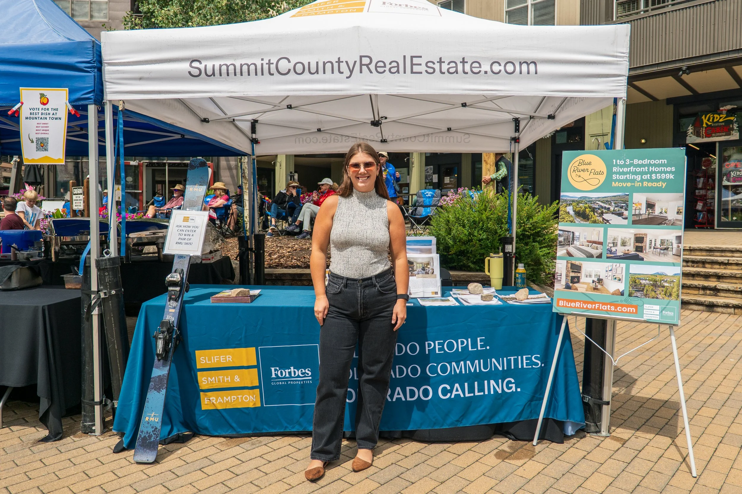 A woman standing in front of a booth at an outdoor event, smiling. The booth has a white banner with 'SummitCountyRealEstate.com' and a blue banner with logos and text promoting real estate services and community information. Other people are sitting