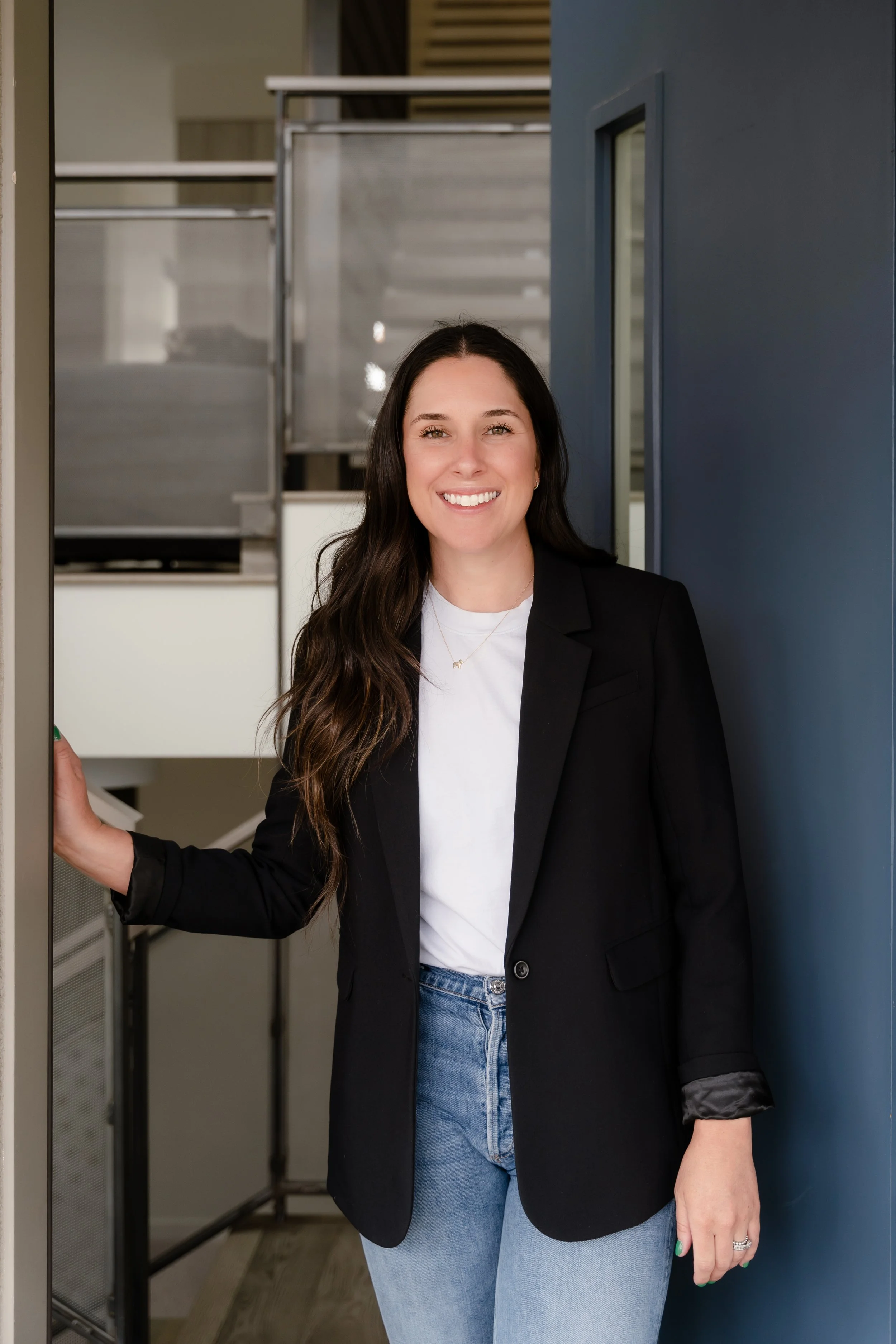 A woman with long dark hair, wearing a black blazer over a white T-shirt and blue jeans, smiling and standing in a doorway with her left hand on the door frame, in an indoor office setting.