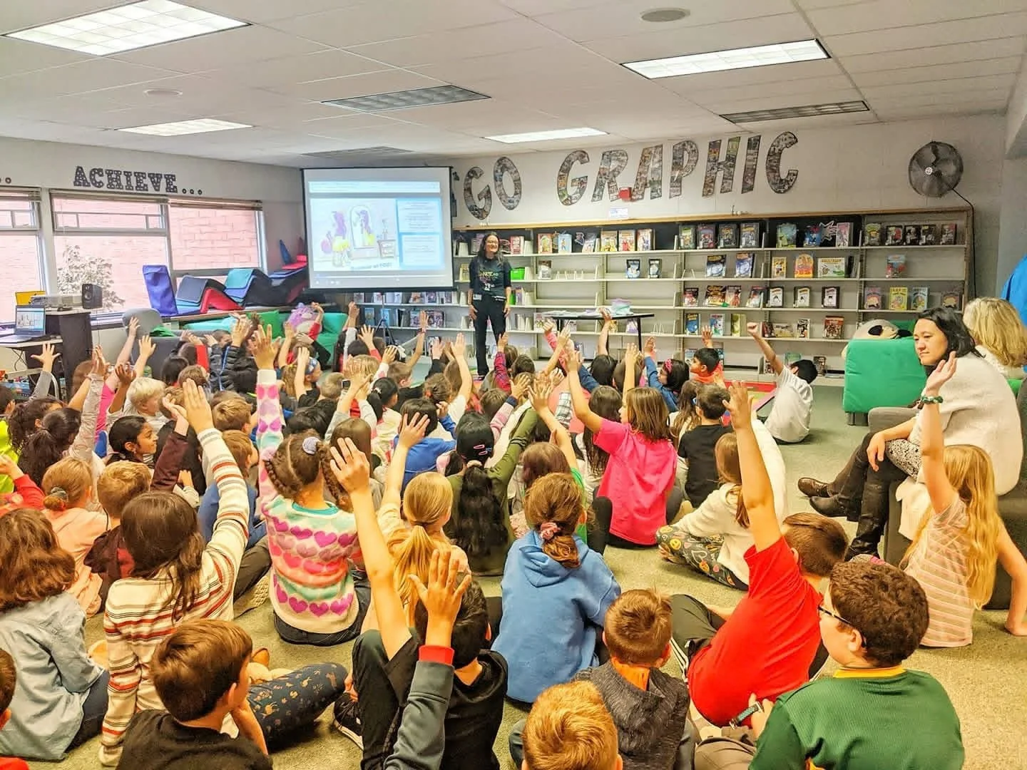 Children raising their hands while a presenter stands beside a projection screen and library shelves.