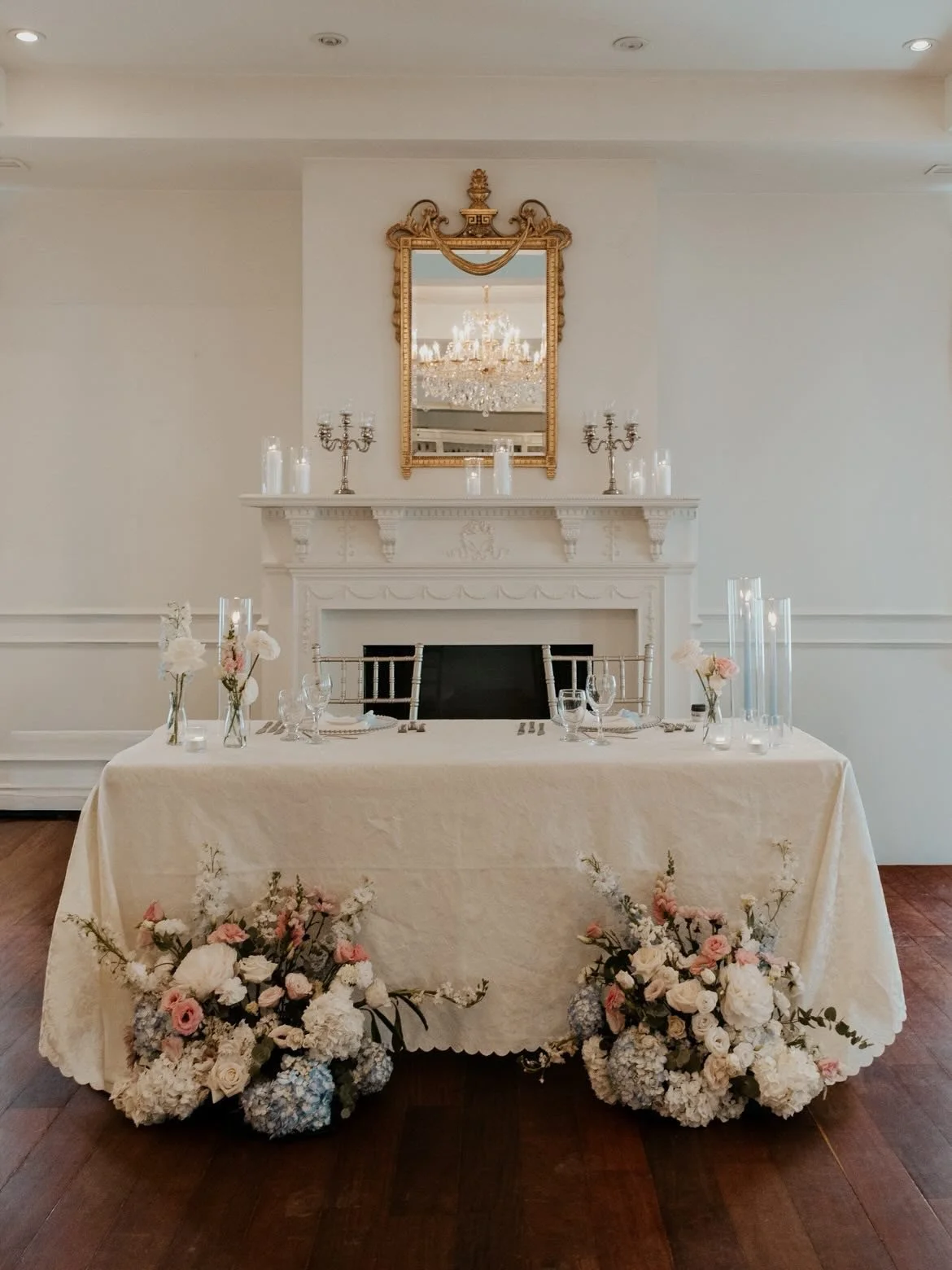 Table de banquet élégante avec nappes blanches, fleurs et bougies, dans une pièce raffinée avec cheminée, miroir doré et chandelier au plafond.