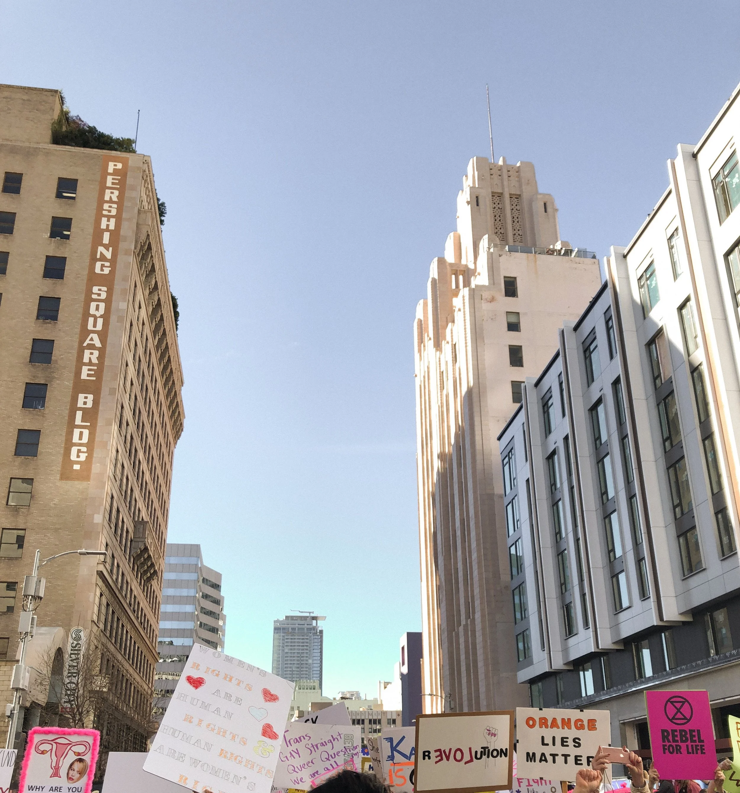 A protest or rally with people holding various signs and banners in a city street, with tall buildings and a clear blue sky overhead.