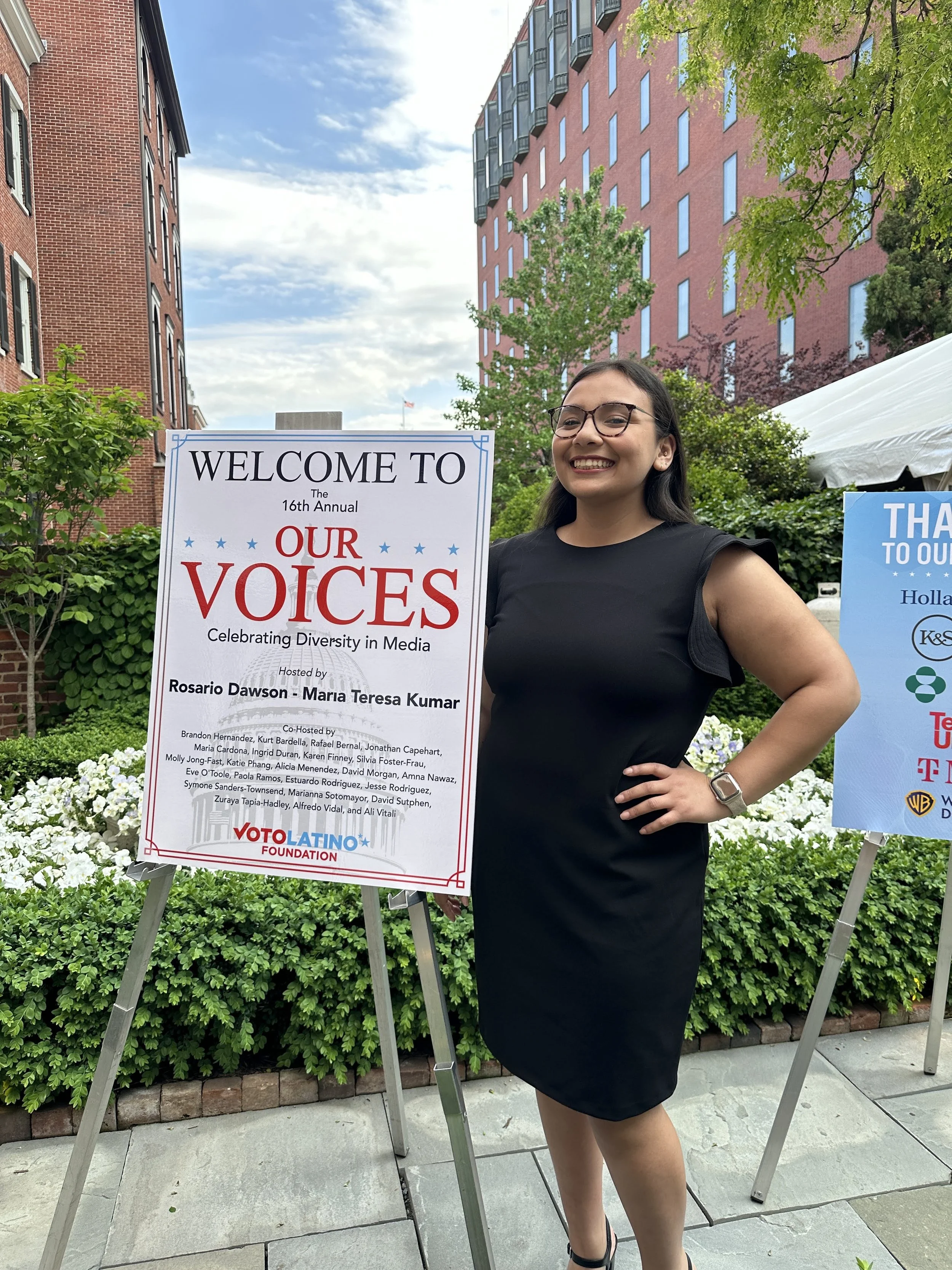 A woman in a black dress and glasses smiling and standing next to two signs in an outdoor urban setting with brick buildings, trees, and bushes. One sign reads 'Welcome to the 16th Annual Our Voices Celebrating Diversity in Media,' and the other appears to thank various sponsors.