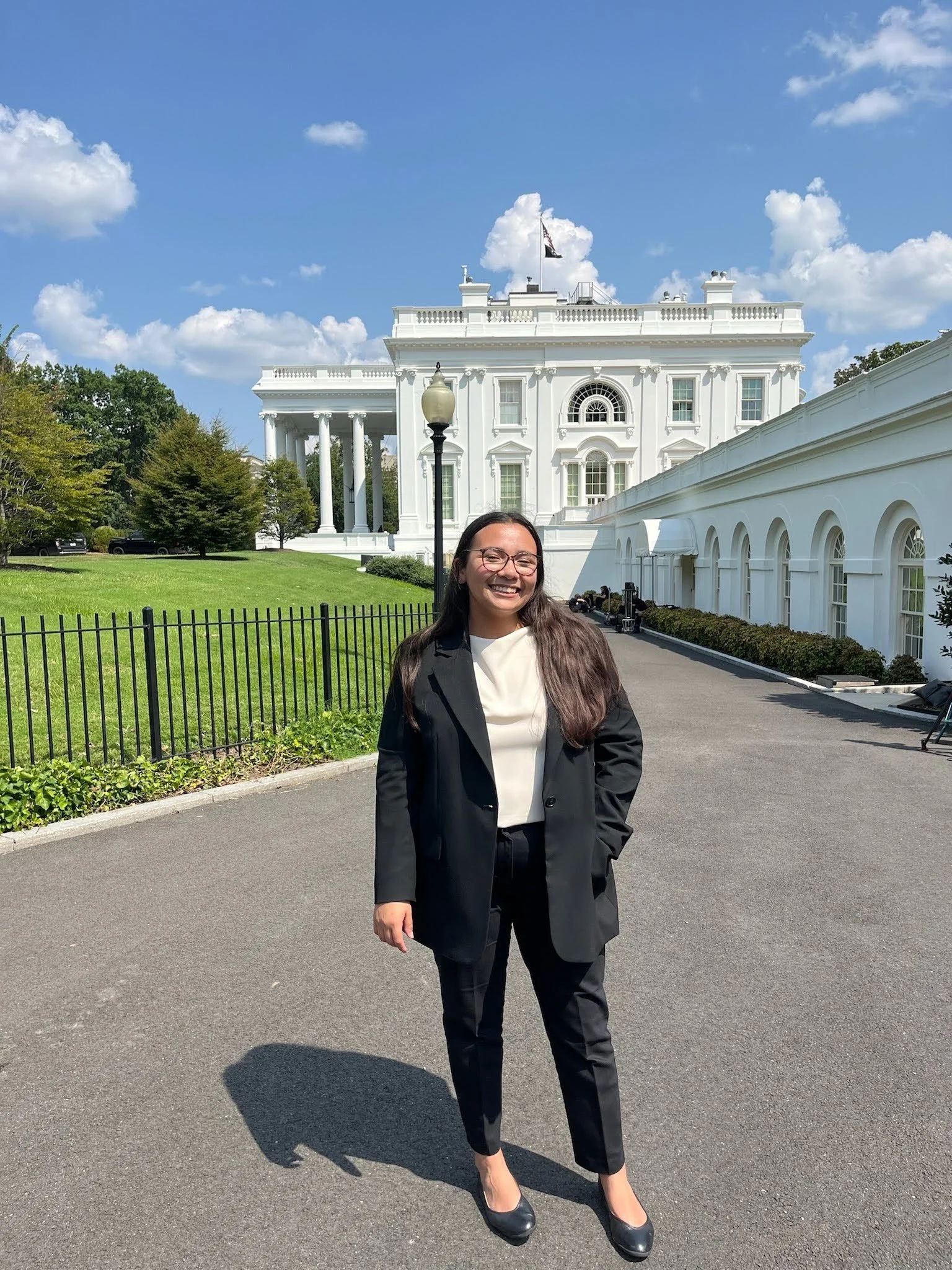 A woman dressed in a black suit with a white top, standing outdoors on a paved area in front of the White House, with a smile on her face and sunglasses on, during daytime under a blue sky with some clouds.
