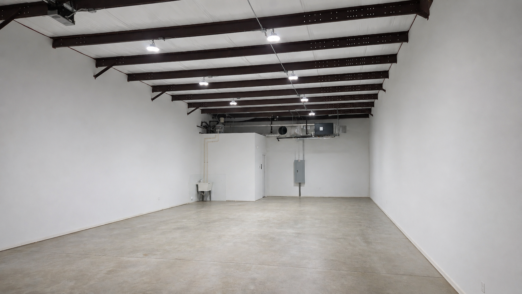 Empty industrial space with white walls, concrete floor, and ceiling beams with lighting fixtures.