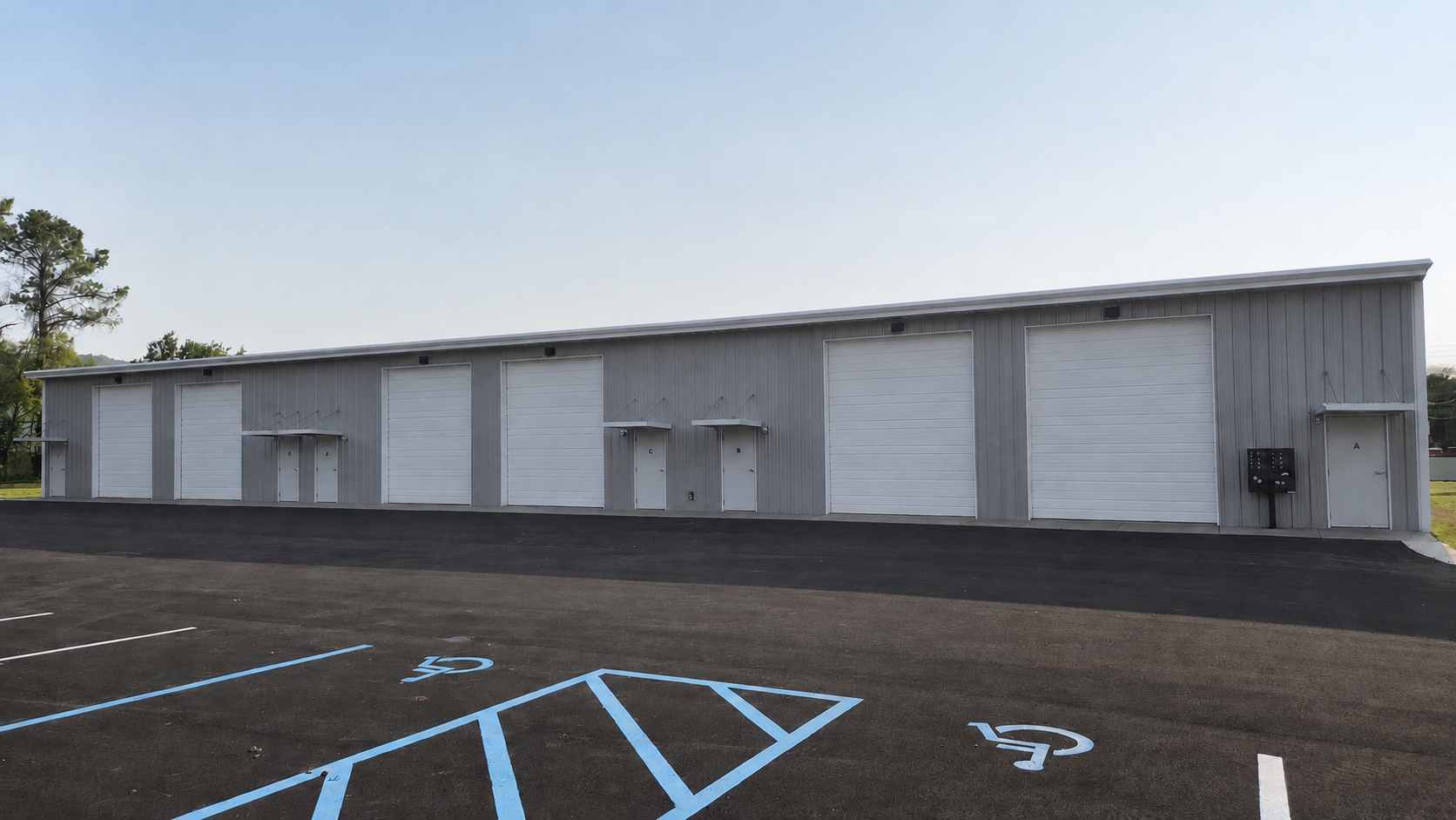 The image shows a large, mostly gray building with several white garage doors and doors marked A, B, and C. There are parking spaces in front, including a marked handicapped spot.