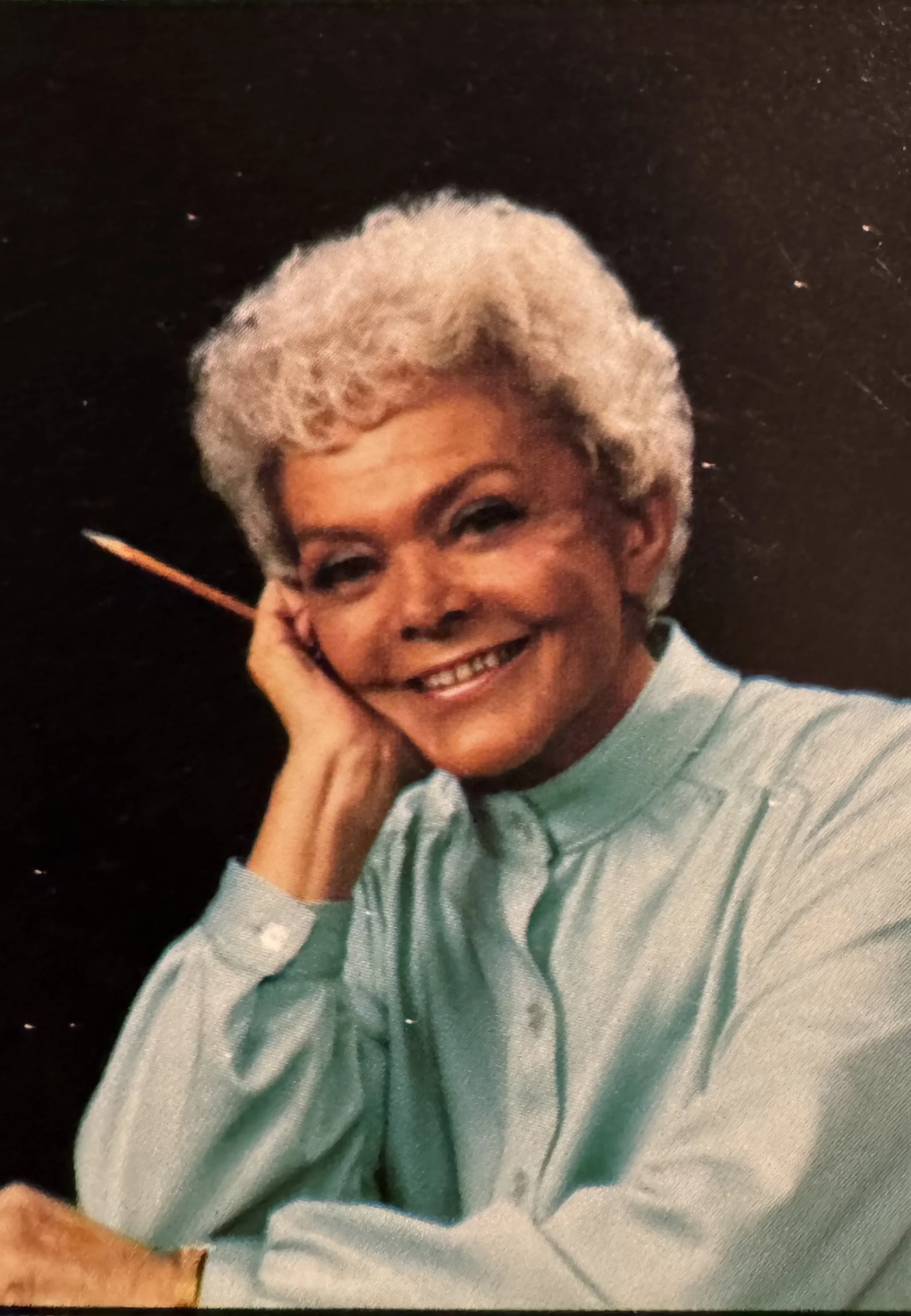 Color portrait of Ethel Barrett, author and storyteller, smiling with short gray hair and resting her head on her hand.