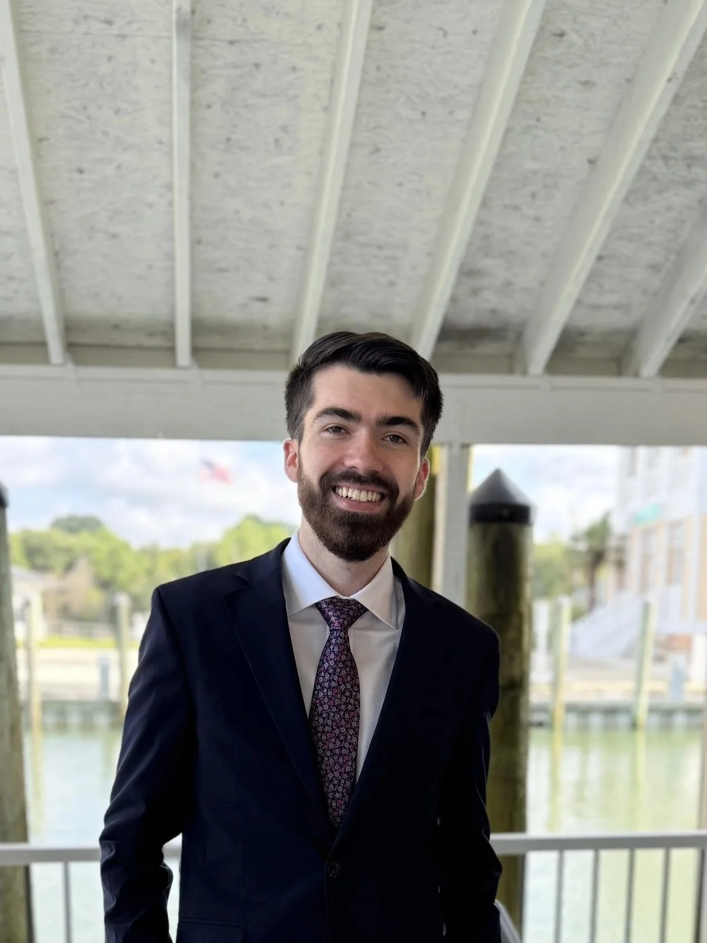 Man in blue suit, white shirt, and patterned tie smiling outdoors beneath white wooden roof with water and bridge in background.