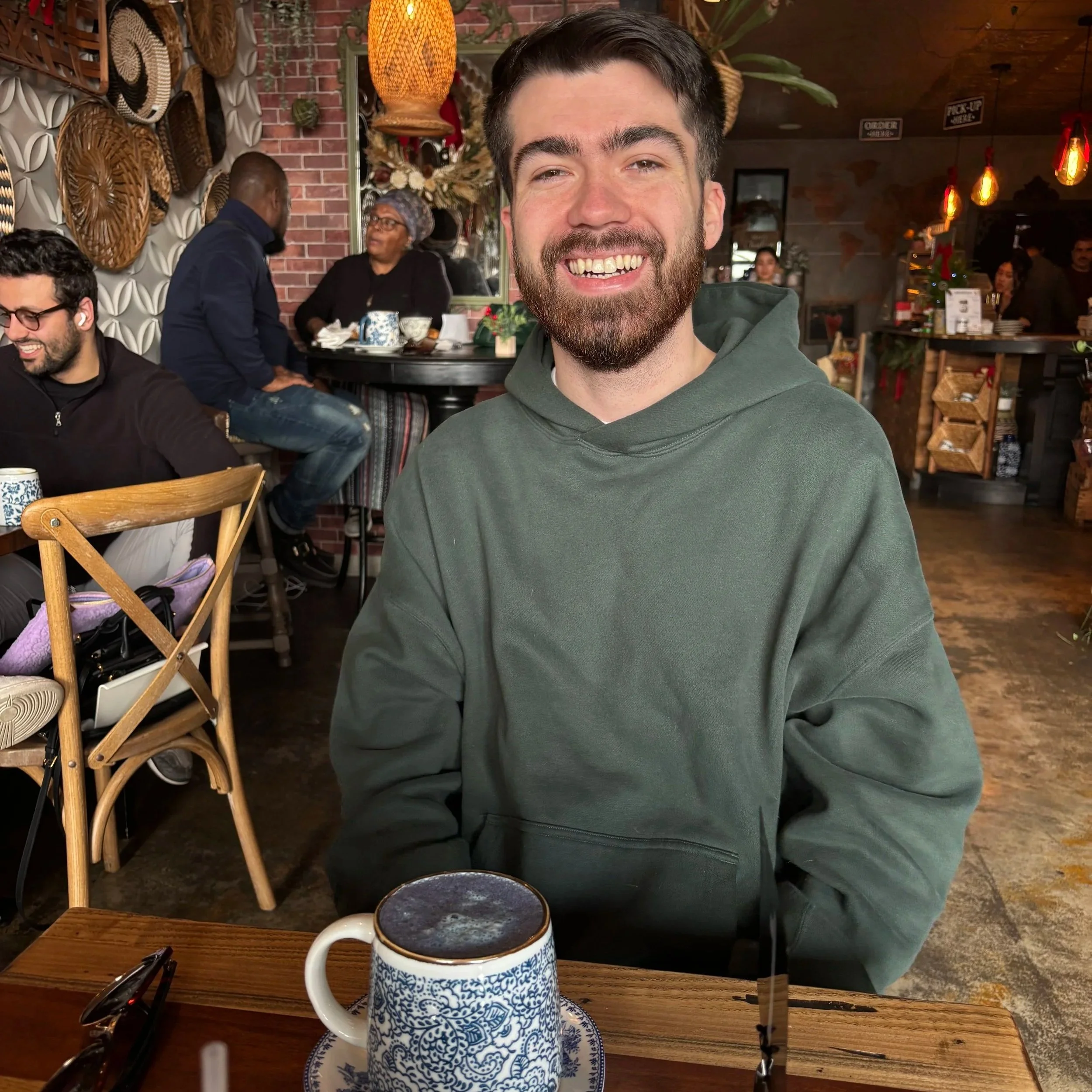 Man with dark hair and beard smiling in cafe, wearing green hoodie. Cafe has warm lighting with hanging pendant lights, decorative wooden baskets on wall, and other people sitting at tables in background.