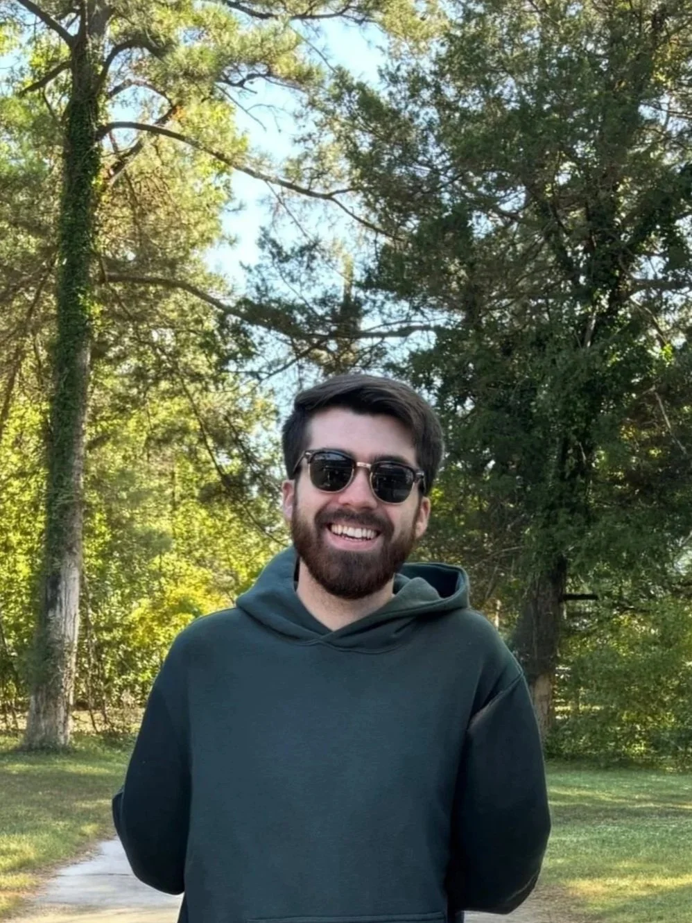 Smiling man with dark hair, beard, and sunglasses standing outdoors on sunny day, surrounded by green trees.