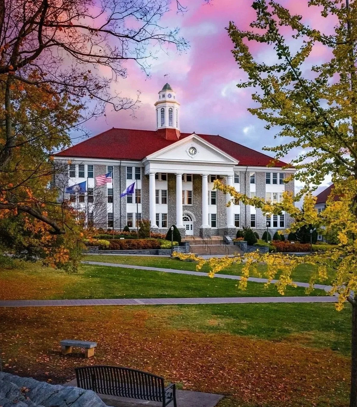 Large, historic building on JMU's campus, with white columns and red roof, framed by trees with changing leaves, under colorful sunset sky.