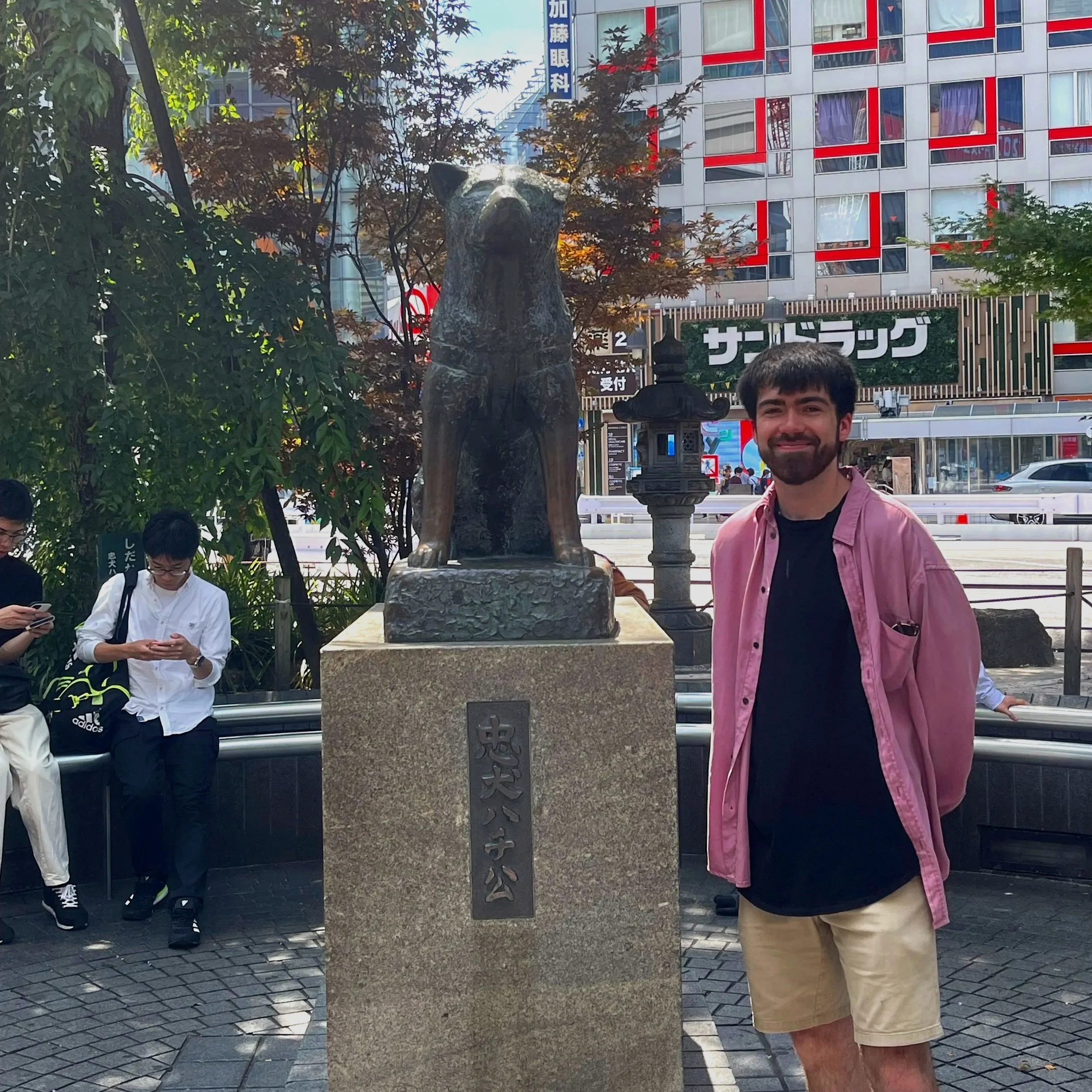 Smiling man with a beard wearing pink shirt and beige shorts standing next to Hachiko stone monument in Japanese urban setting with tall buildings, trees, and young people in background.