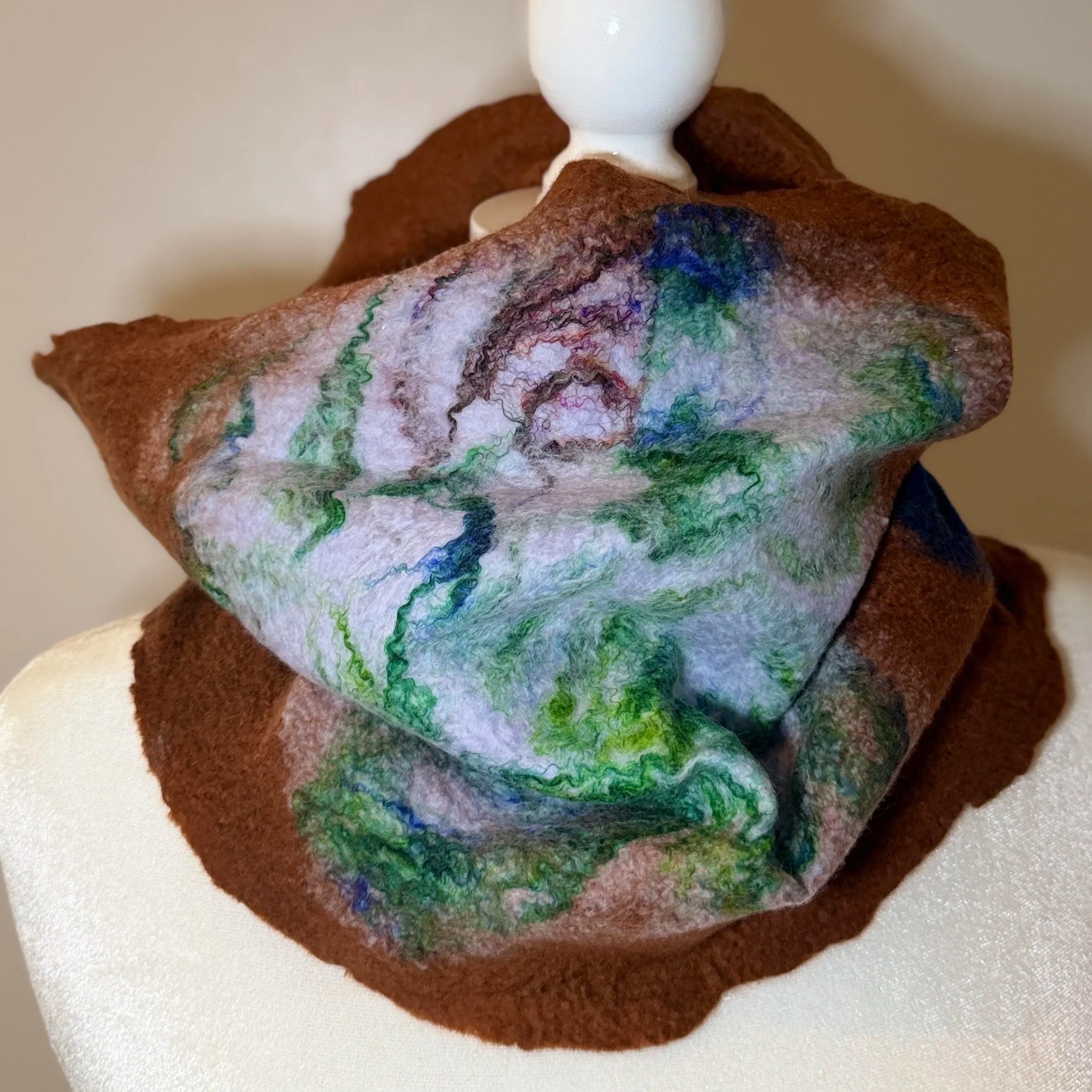 A felted wool art sculpture of a mountain landscape with colorful abstract lines, a white cloud on top, and a brown base, placed on a white surface.