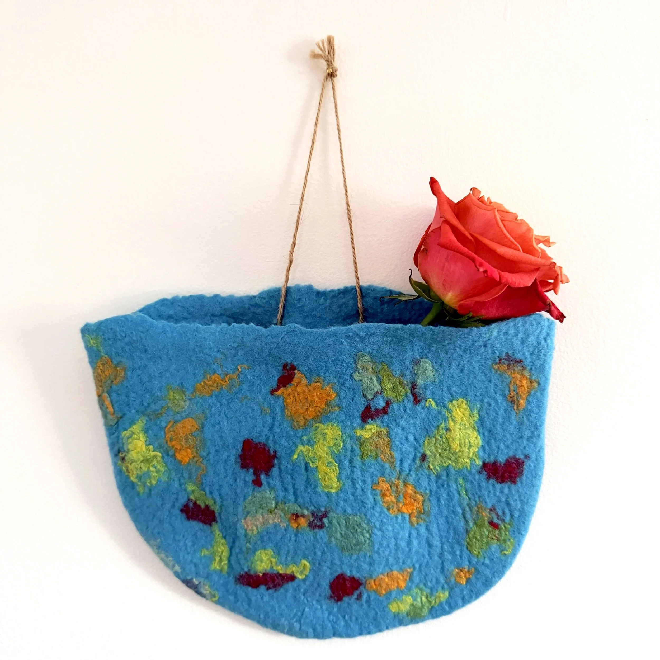 A colorful, felted art piece with a blue base, decorated with multicolored wool roving, a red-pink rose, and a hanging rope.