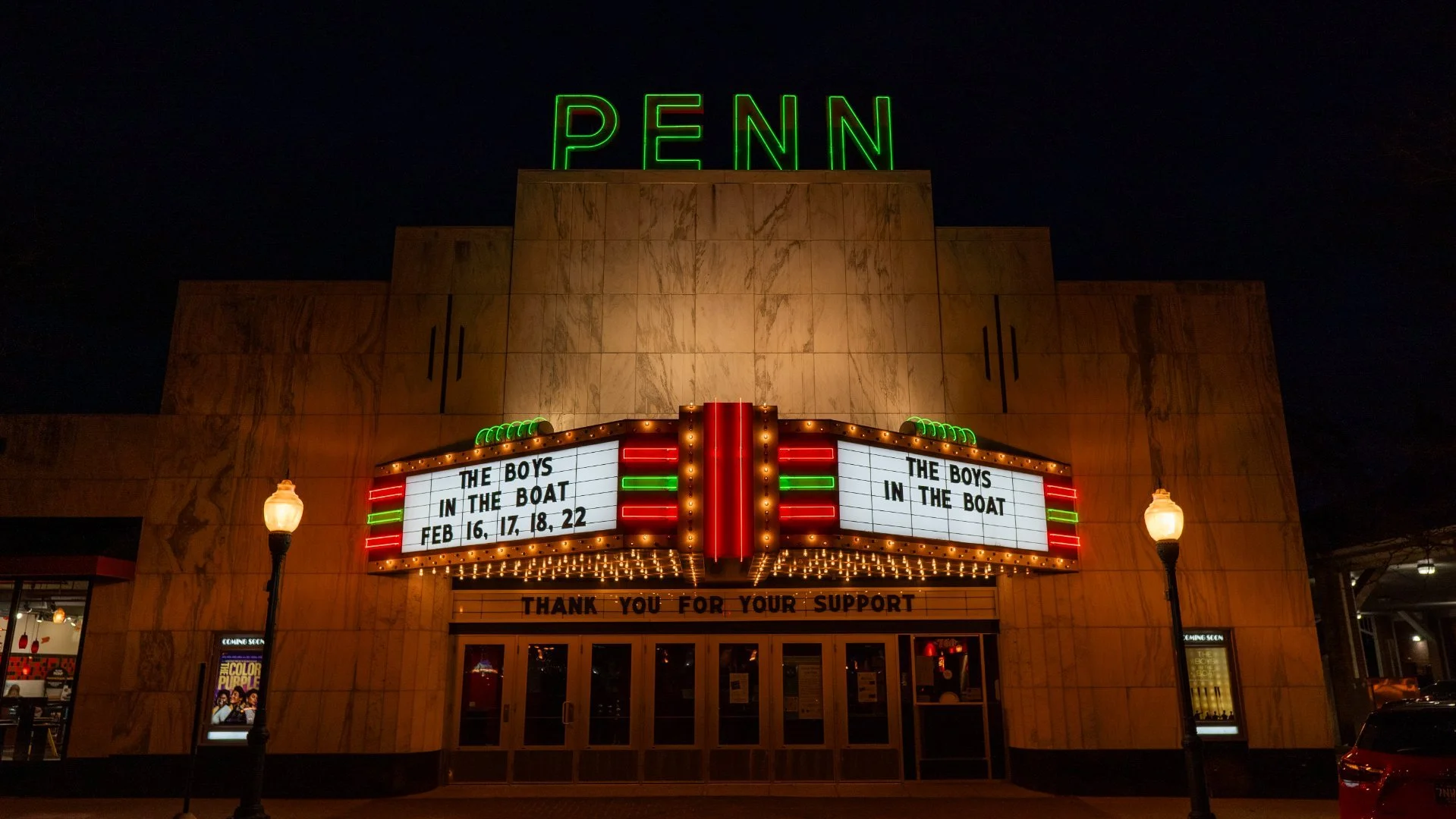 Marquee sign outside of a theater at night, advertising a show called "The Boys in the Boat" with dates February 16, 17, 18, and 22. The building features large neon letters spelling "PENN" at the top, and the theater's entrance is flanked by vintage-style lampposts.
