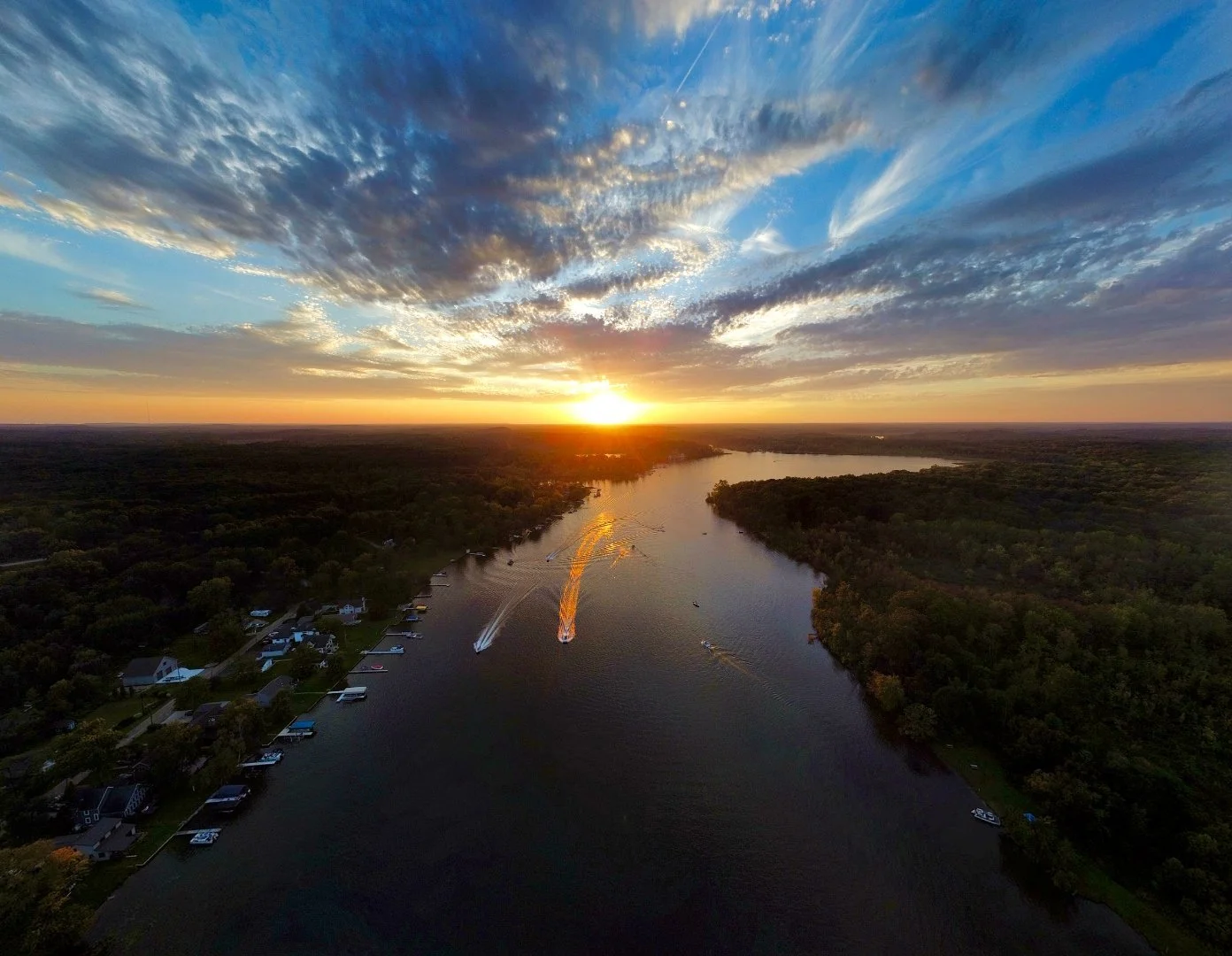 An aerial view of a wide river reflecting the sunset, with boats on the water, surrounded by lush green forests and a small community along the shoreline, under a vibrant sky with clouds at sunset.
