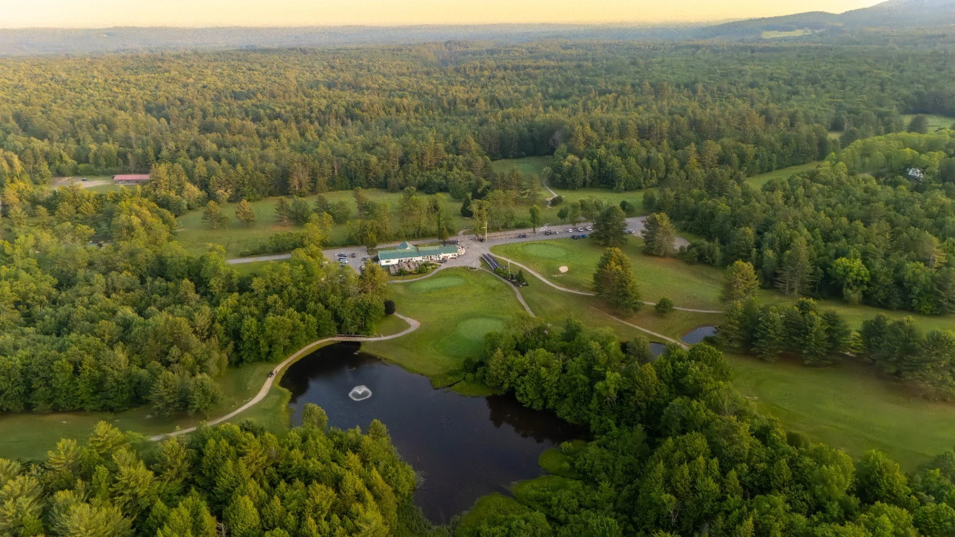 An aerial view of a golf course surrounded by dense trees and a large parking lot with a building nearby, a pond with a fountain, walking paths, and a few golf carts.