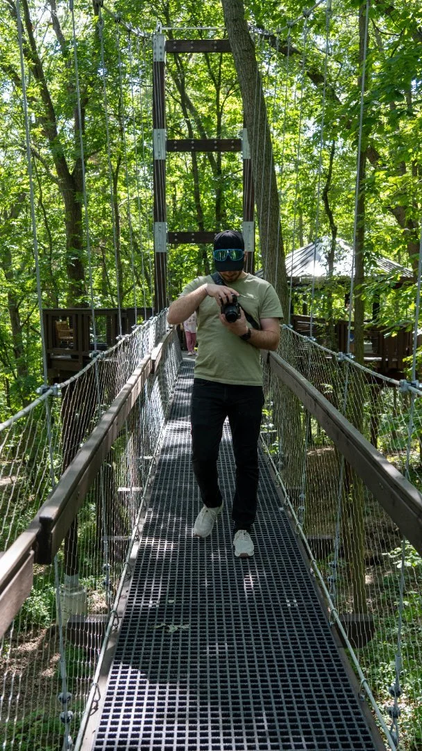 A person taking a photo with a camera on a hanging bridge in a green forest.