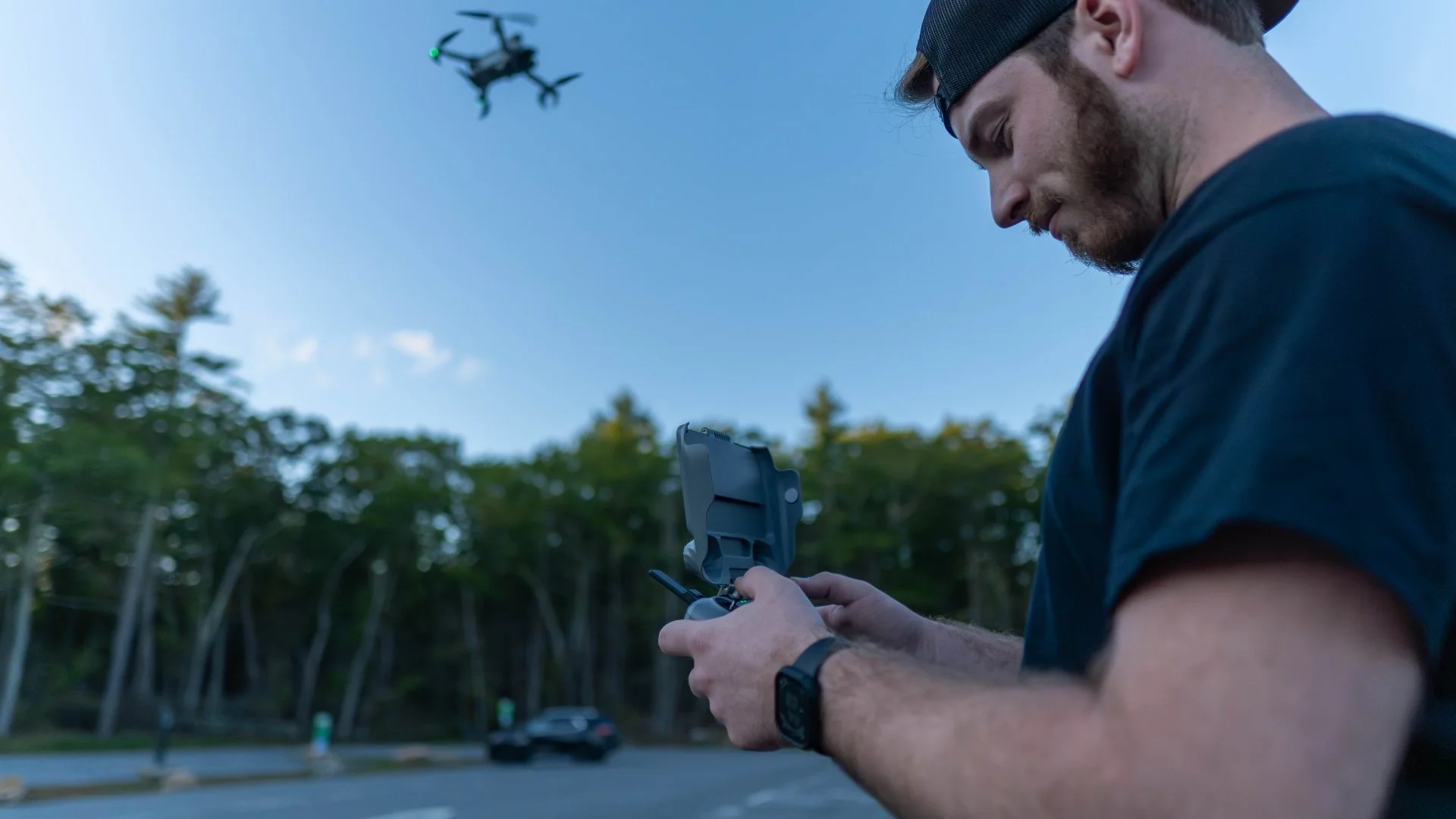 A man standing outdoors at dusk, looking down at a remote control with a drone flying overhead.