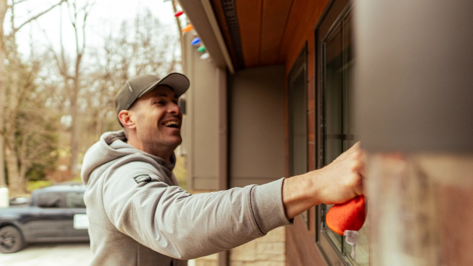 A man smiling as he reaches into a mailbox on a porch, wearing a gray hoodie and a gray cap, with trees and parked cars in the background.