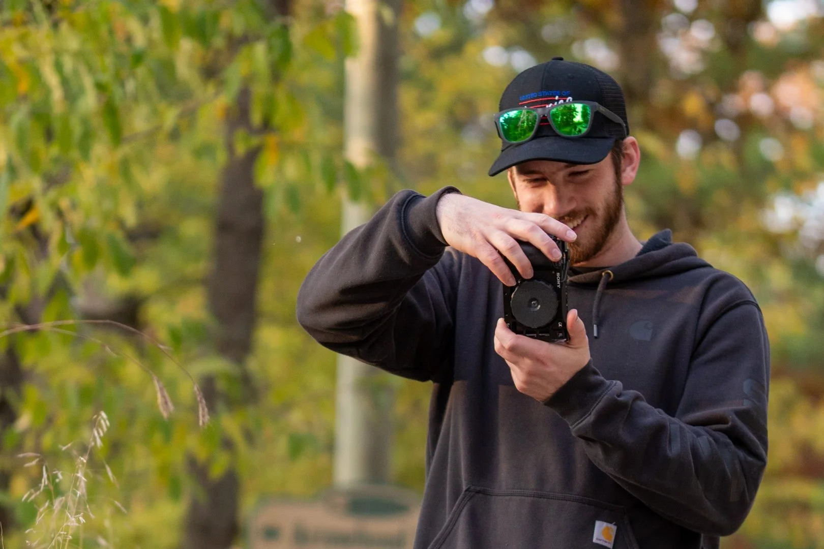 A young man with a beard, wearing a black baseball cap with green sunglasses attached, and a black hoodie, smiling as he takes a picture with a camera in an outdoor setting with trees and foliage in the background.