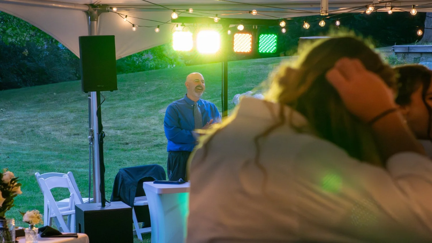 A man in a blue shirt and patterned tie speaking at an outdoor event under a white canopy, with string lights and a colorful lighting setup, while guests are seated in front of him.