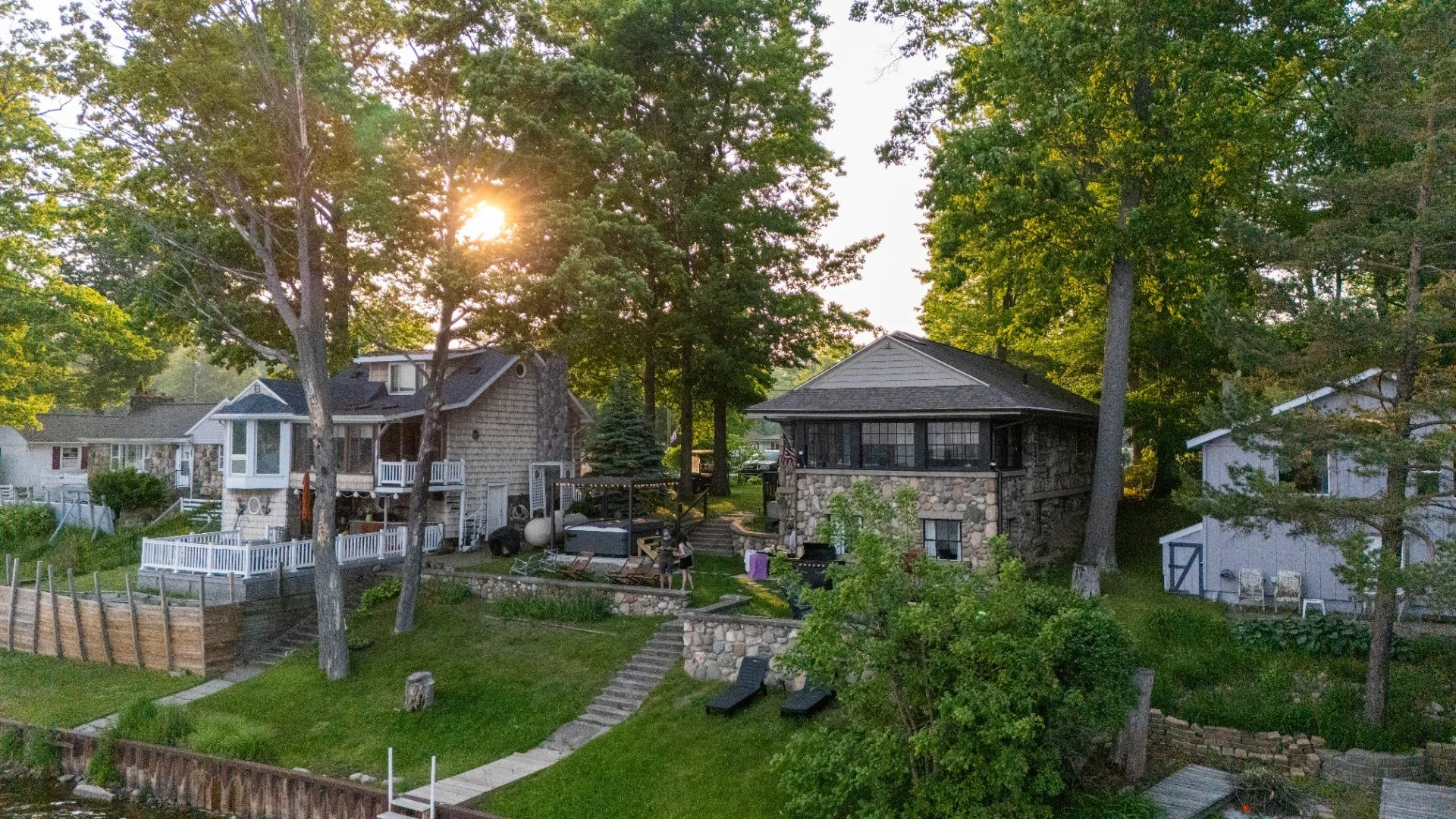 A backyard scene with two houses, trees, and a garden during sunset. One house has a porch, the other has a stone facade. There are steps, outdoor furniture, and yard decorations.