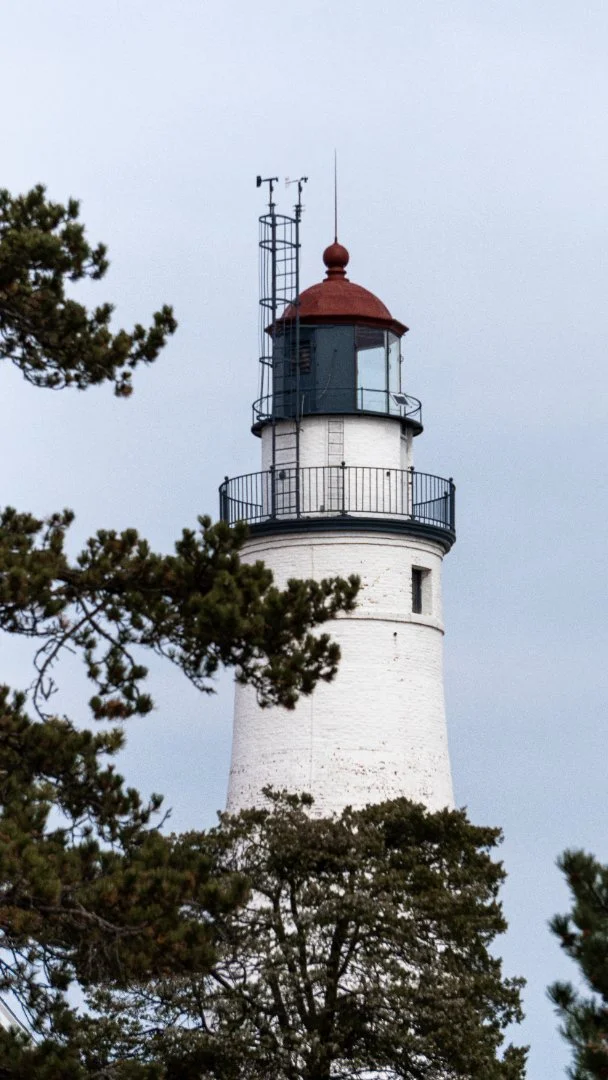 A white lighthouse with a red roof, black railing around the top platform, and antennas, partially obscured by trees against a cloudy sky. - Taken by Dillon Morby of D Morby Media, a local photographer working in South East Michigan