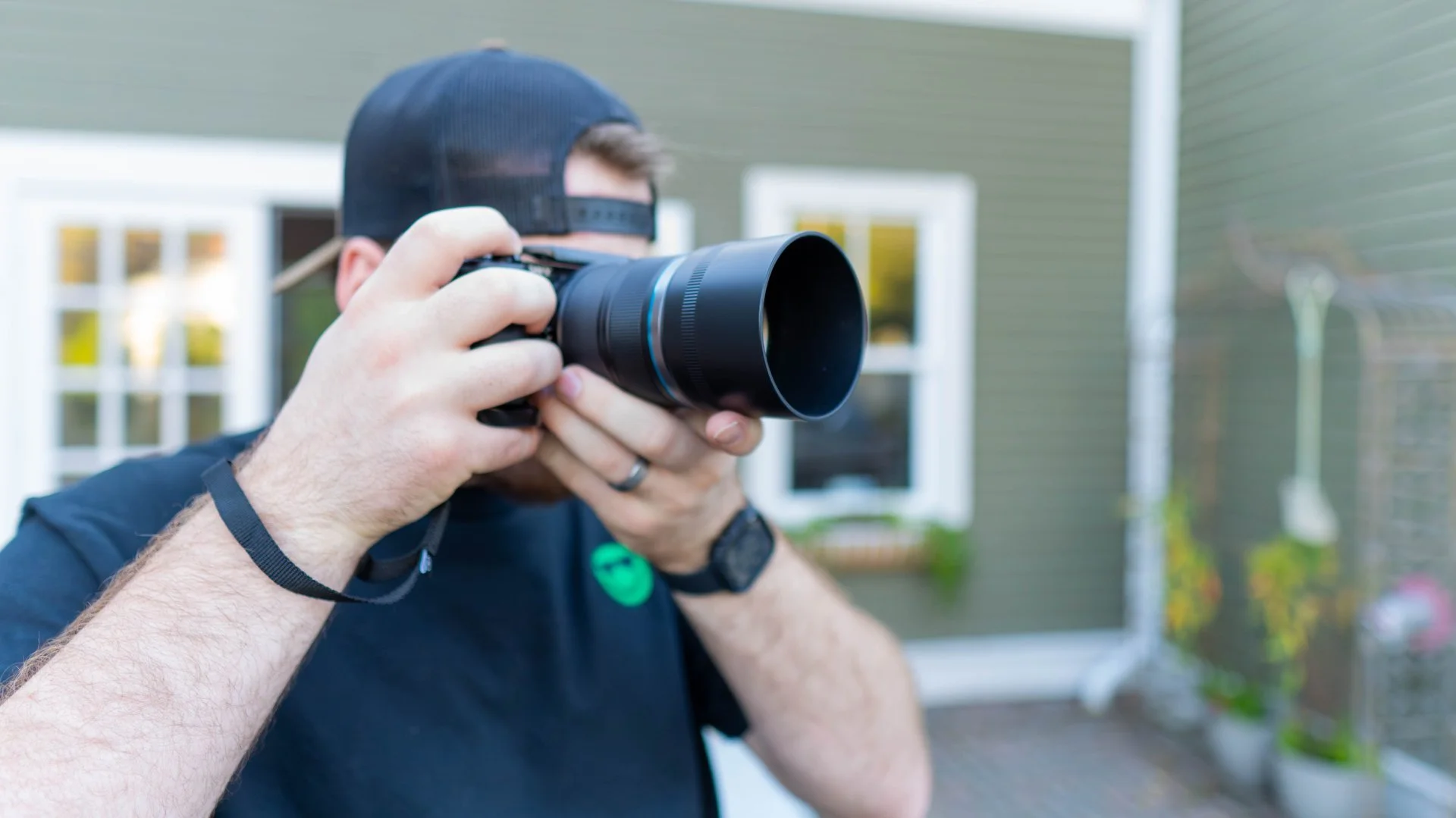 A man wearing a black cap and t-shirt is holding a camera up to his eye, taking a photograph outside in front of a house with green siding and white trim.