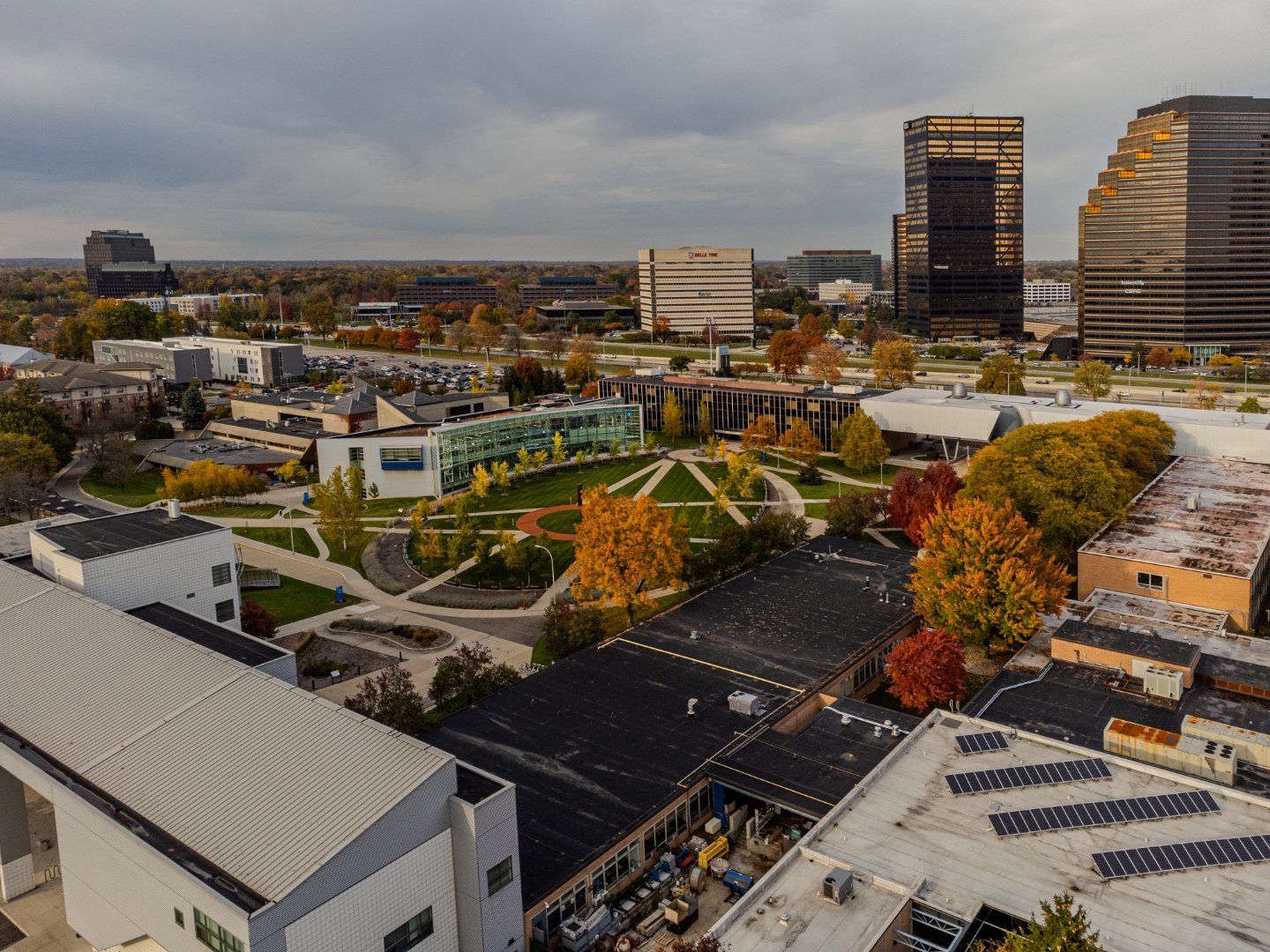 A cityscape featuring modern buildings, a park with trees in autumn colors, and a cloudy sky.
