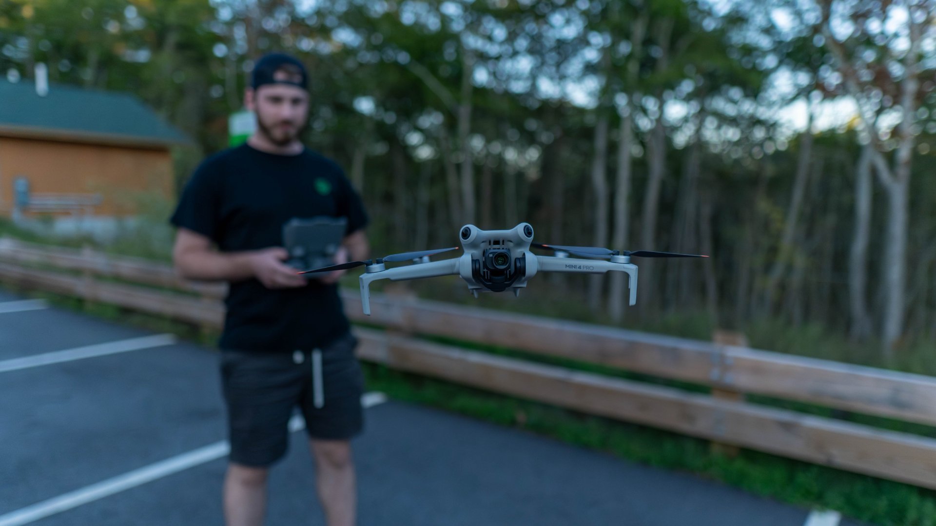 A man in black clothing and a black cap is flying a drone outdoors near a wooded area with trees and a wooden fence, holding a remote control device.