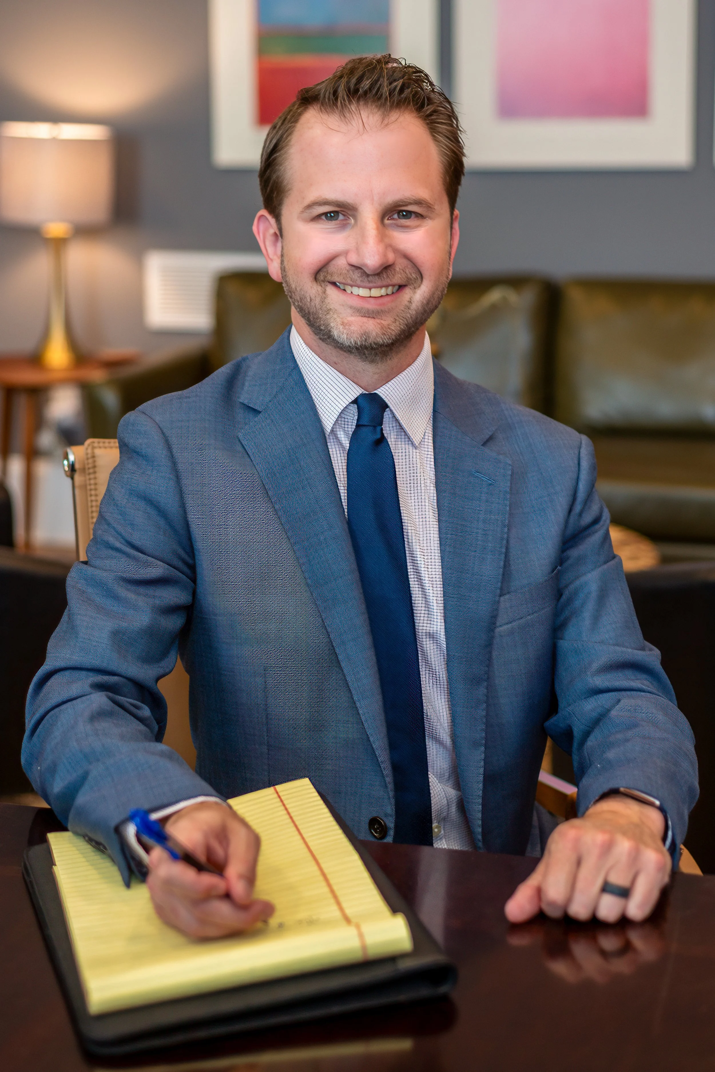A man in a gray suit and blue tie sitting at a table, smiling, with a yellow notepad and pen in front of him. The background shows a gray wall with framed colorful artwork and a table lamp.