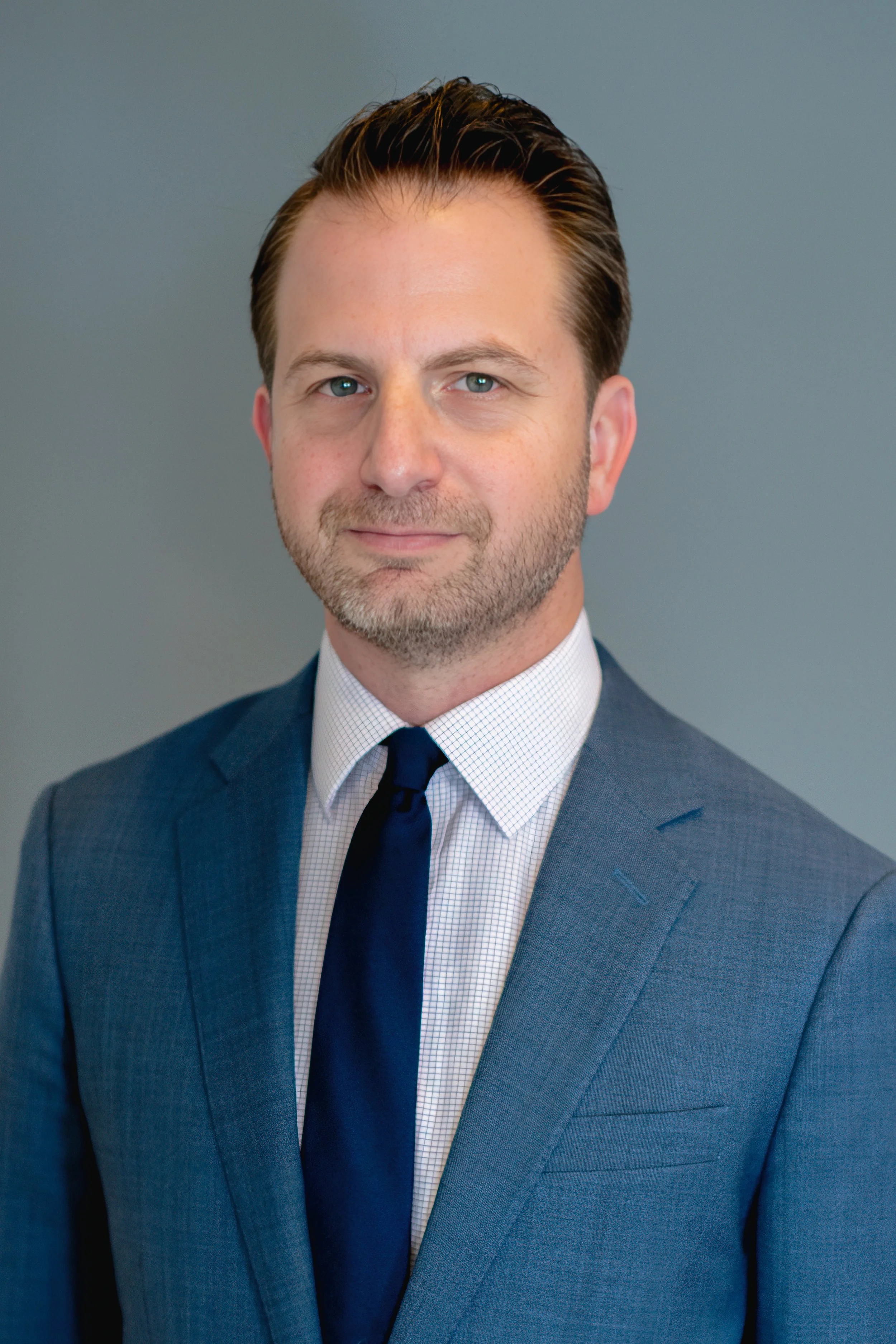 Headshot of a middle-aged man with light skin, blue eyes, brown hair, and a beard, wearing a navy suit, white checkered shirt, and navy tie, against a plain blue background.