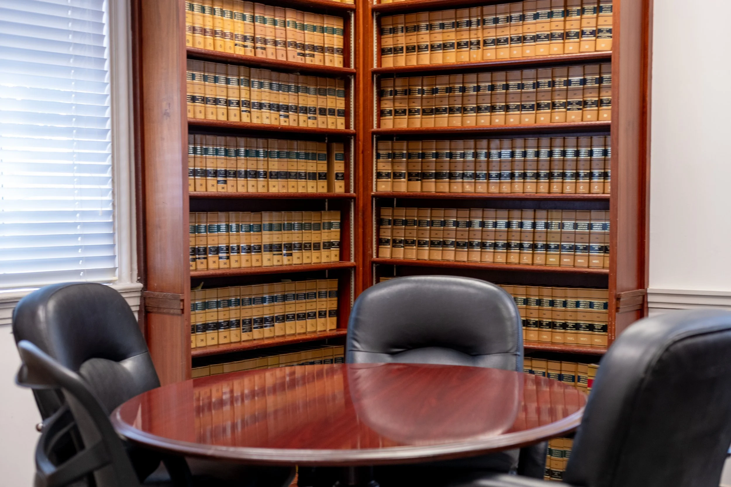 A conference room with a large wooden table surrounded by black chairs. Behind the table, there is a tall wooden bookshelf filled with legal books or volumes, and a window with white blinds partially open.
