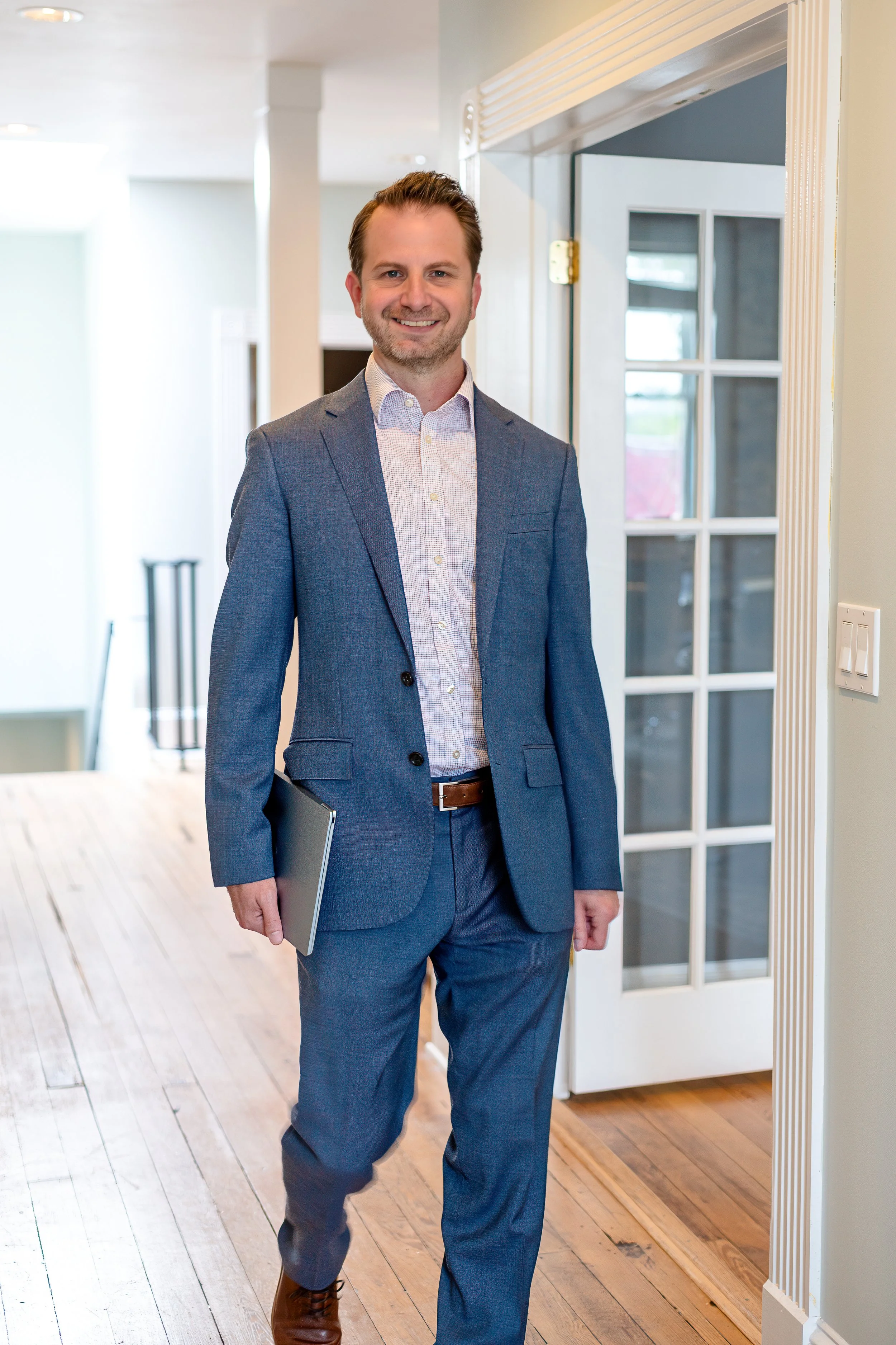 A man wearing a blue suit and white shirt holding a laptop and smiling, standing in a bright room with wooden floors and white walls.
