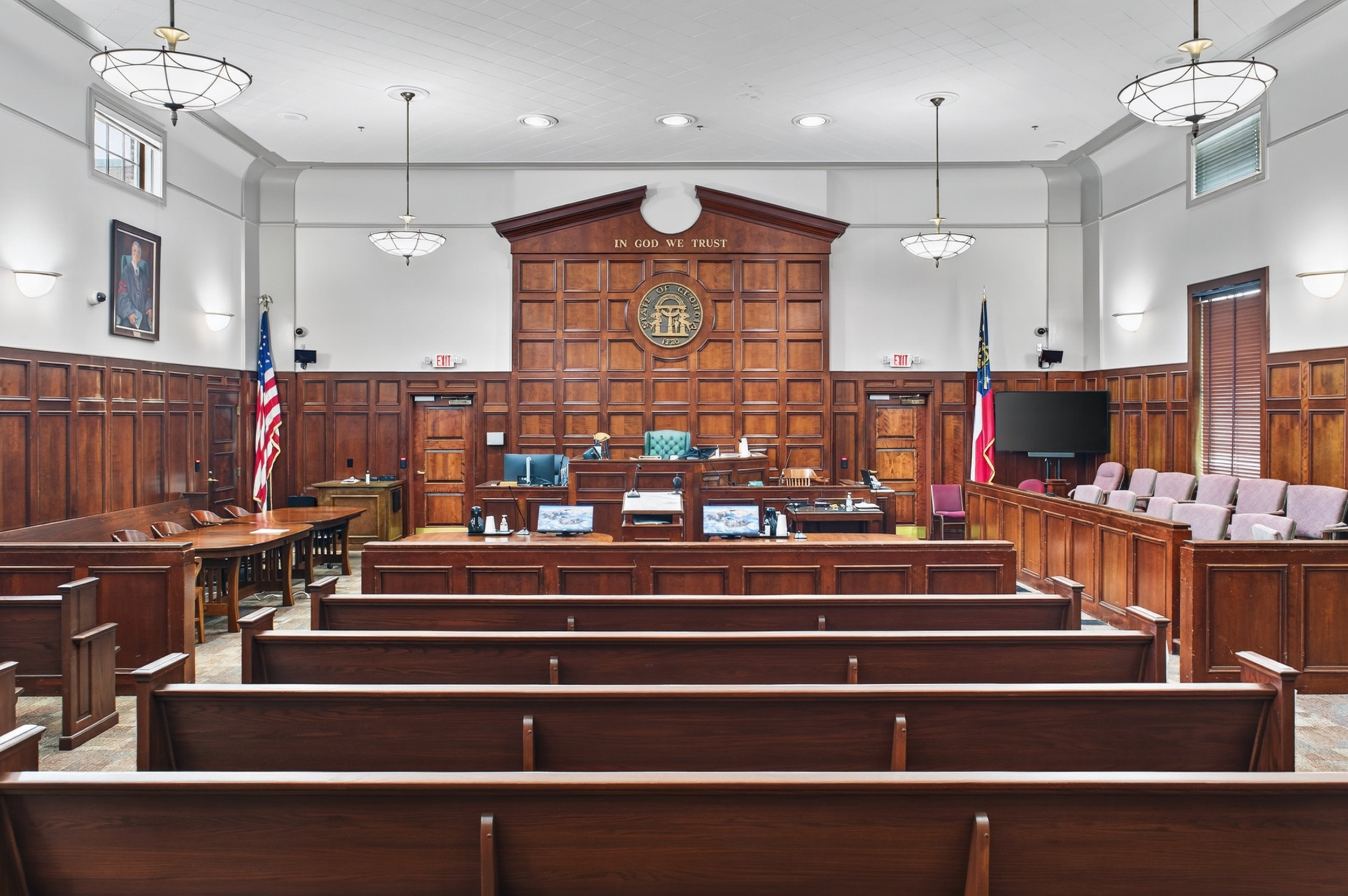 A courtroom with wooden paneling, a judge's bench in the center, portraits on the walls, a large clock, and flags, with chairs for spectators and legal staff.