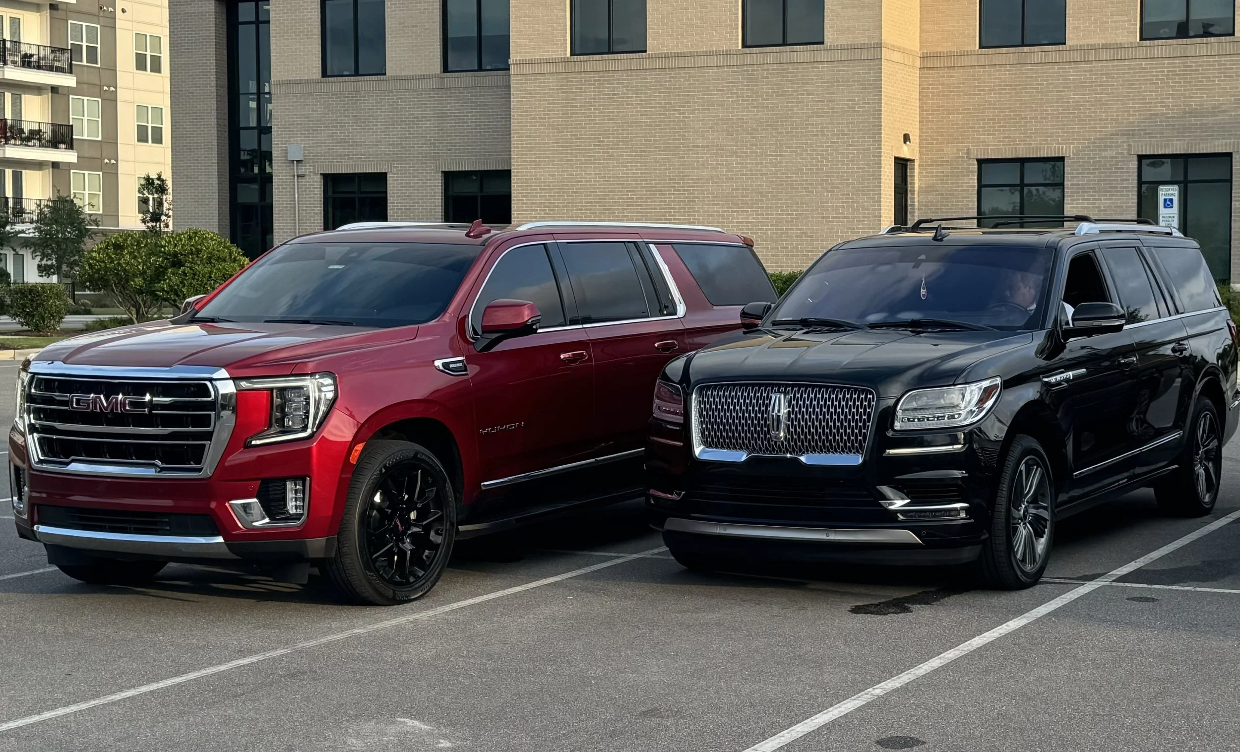 A red GMC SUV and a black Lincoln SUV parked side by side in a parking lot in front of a modern brick building.