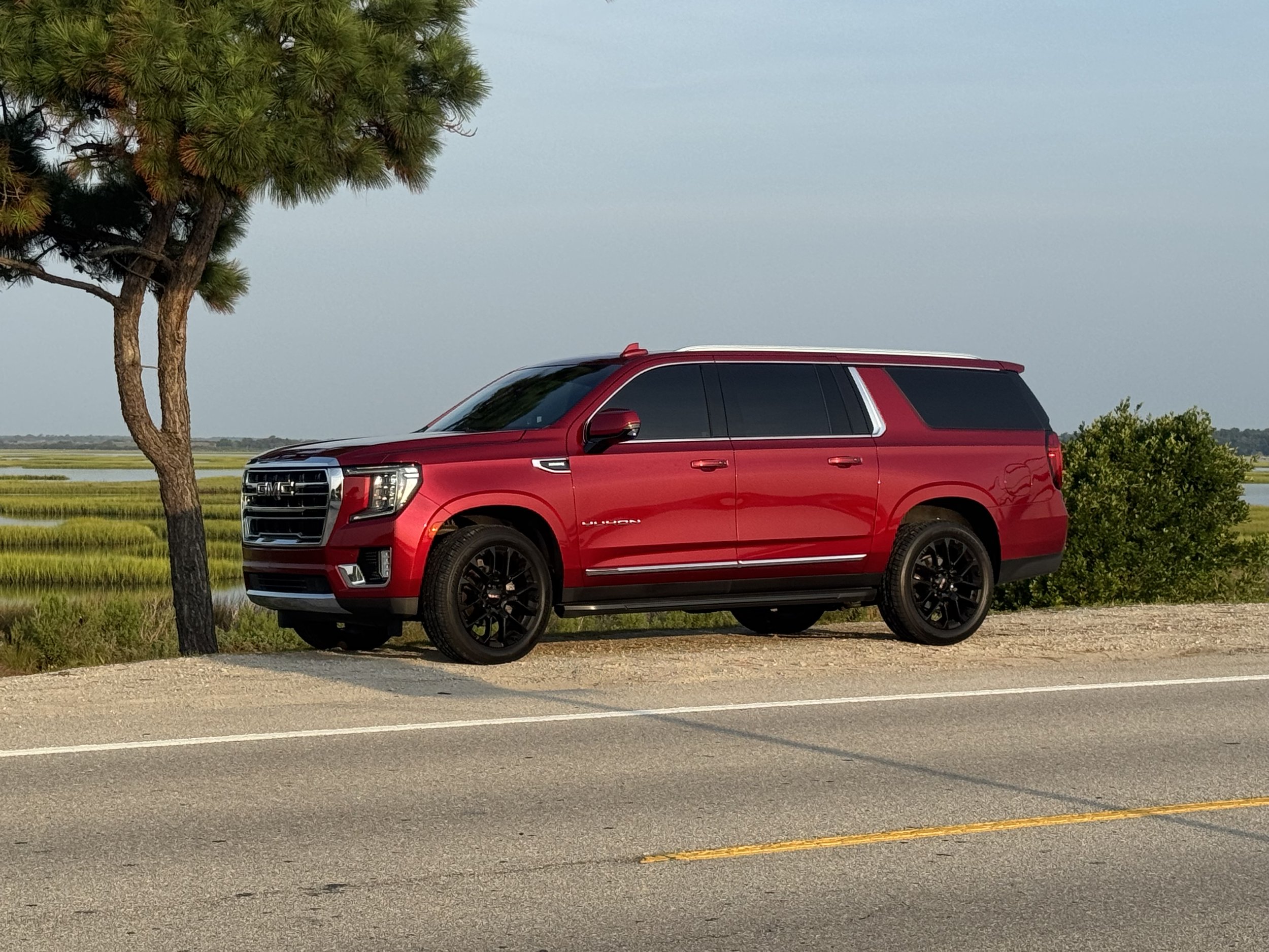 Red GMC SUV parked on the side of a rural road with green fields, a large tree, and a cloudy sky in the background.