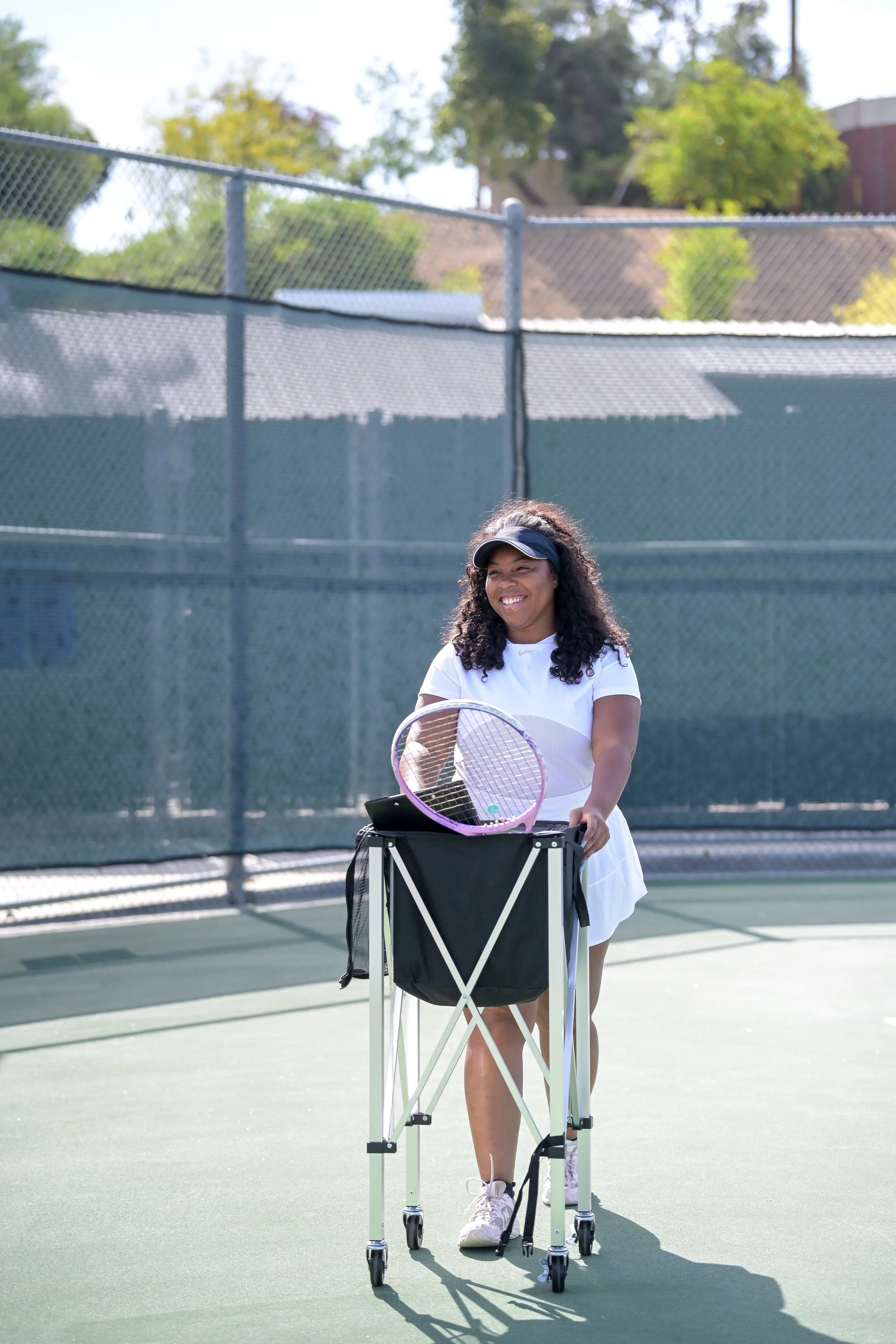 A woman with curly hair, wearing a visor, white tennis outfit, and sneakers, standing on a tennis court with a tennis racket, using a walker with a tray carrying tennis balls.